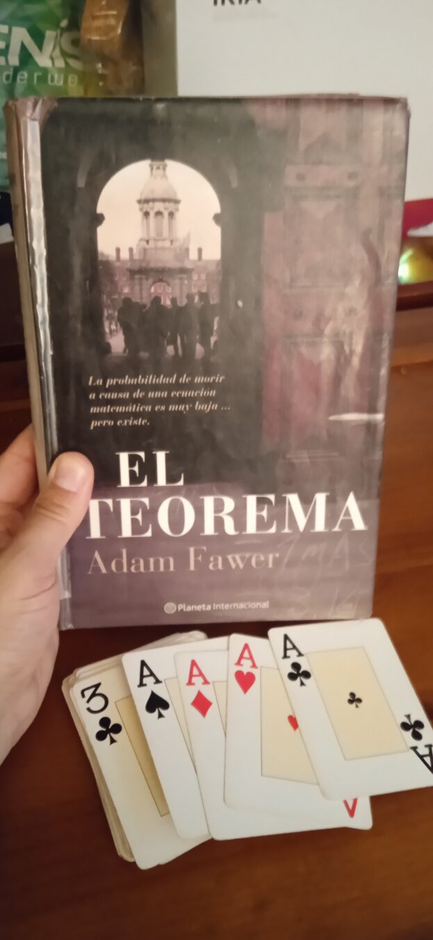 Se ve la portada del libro El teorema de Adam Fawer sujetada por una mano y delante un mazo de cartas mostrando los 4 ases de una baraja de póker