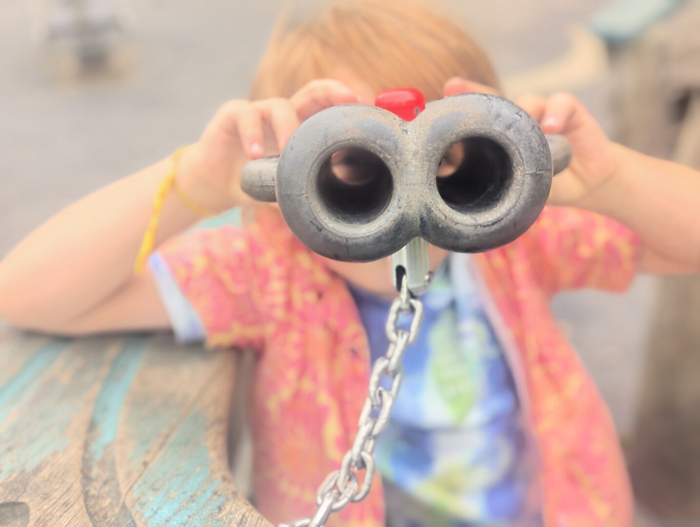 A boy wearing a colourful shirt is holding up a pair of toy binoculars to his eyes, which causes him to be out of focus because the end of the binoculars are the focus.