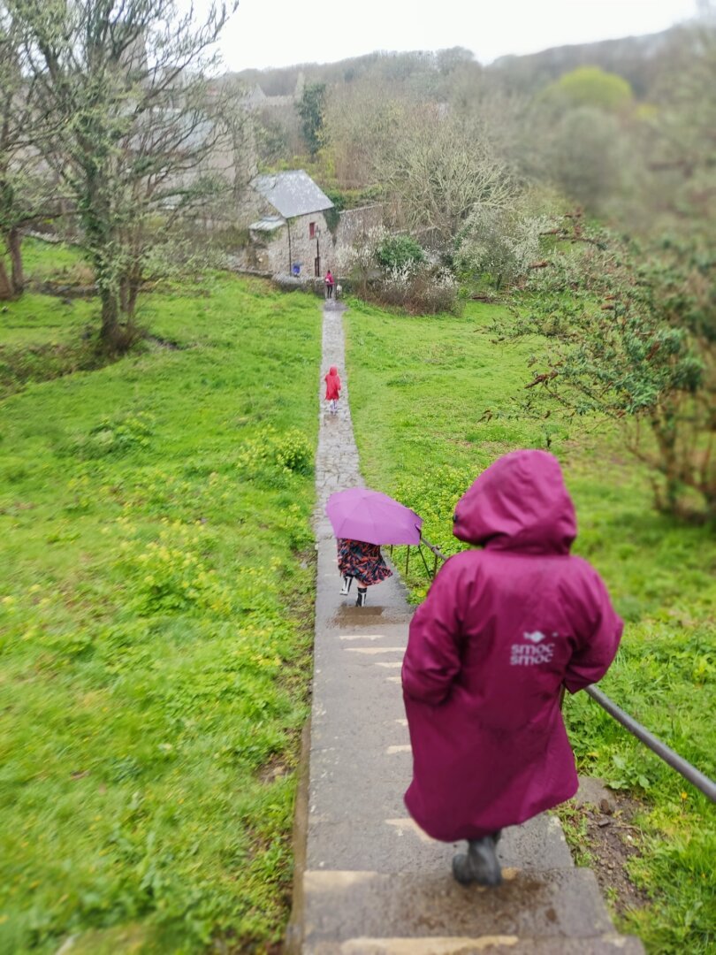 An adult and two children walking down a path with grass and trees on either side. They face away from the camera and are wearing raincoats