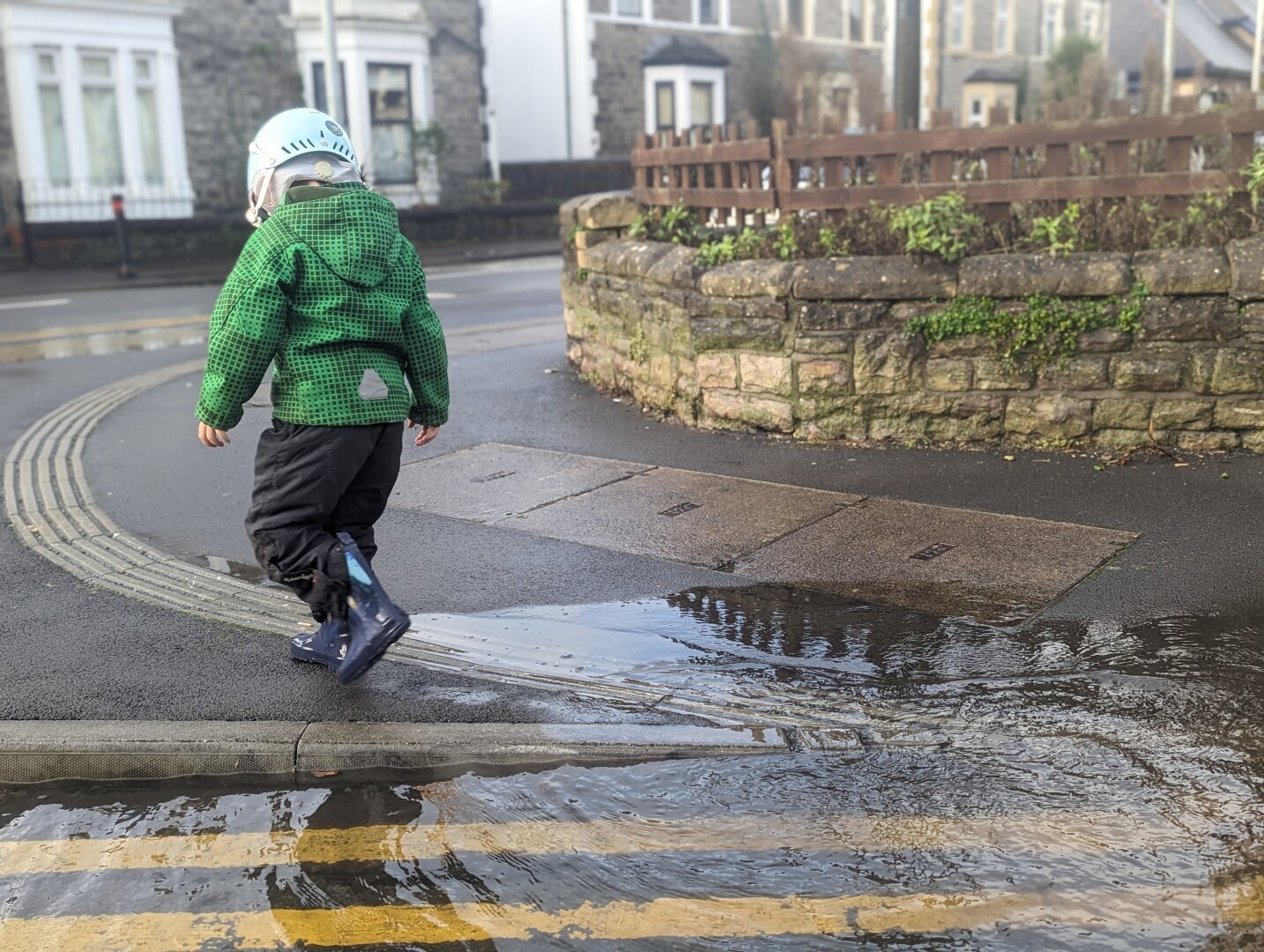 A boy in wellies and a bike helmet splashes through a puddle by some double yellow lines on the street.