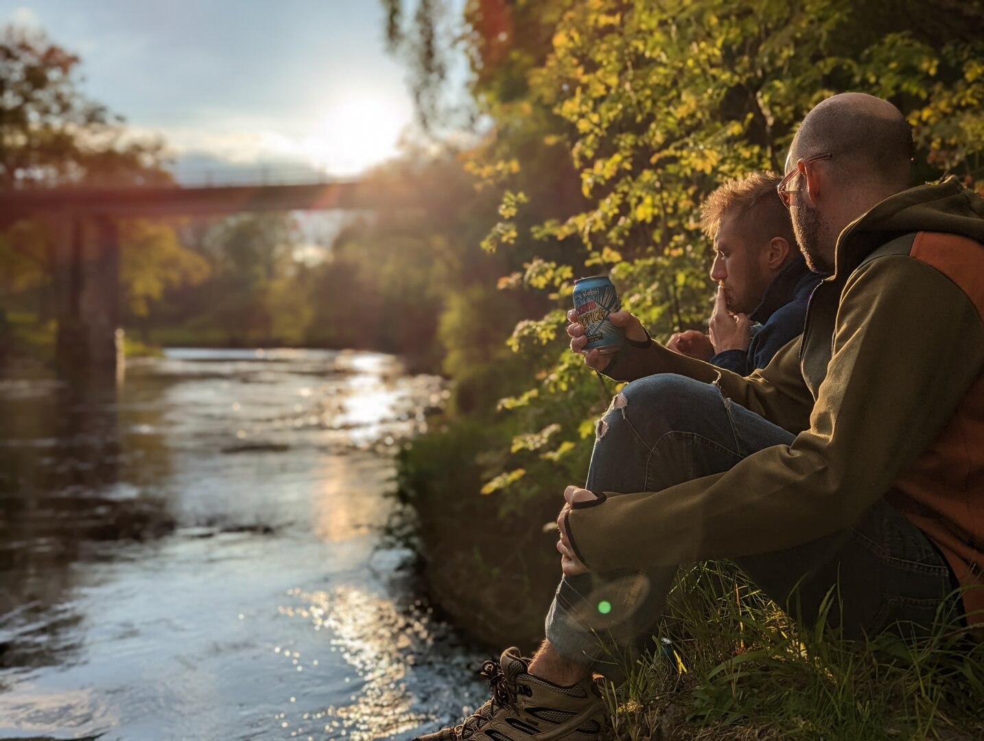 Two men sit on the riverbank at sunset drinking beer. The riverbank is wooded and there is a bridge in the distance.