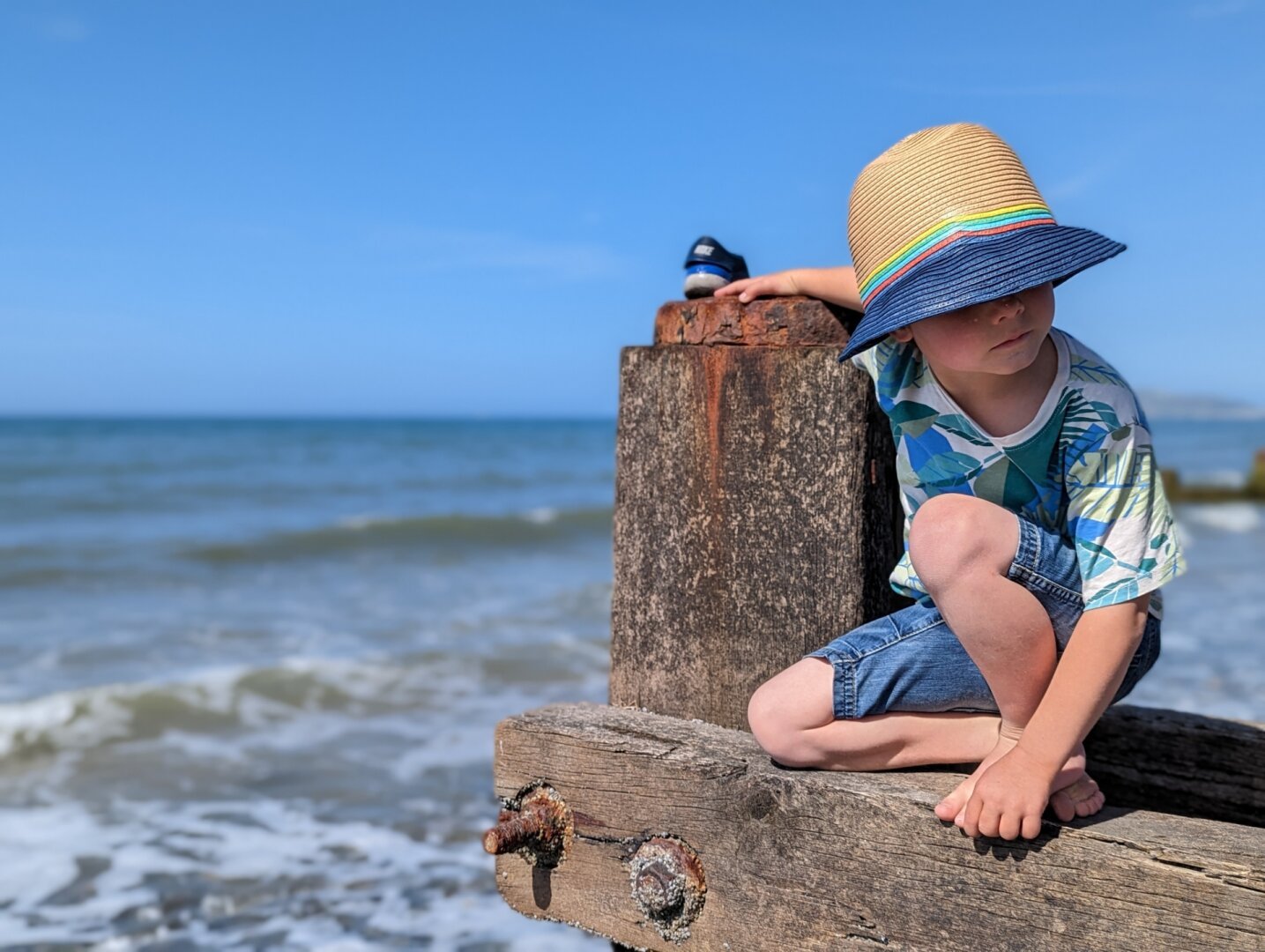 A young boy wearing a hat is sitting on the end of a beach groin. The sea and sky forms the background