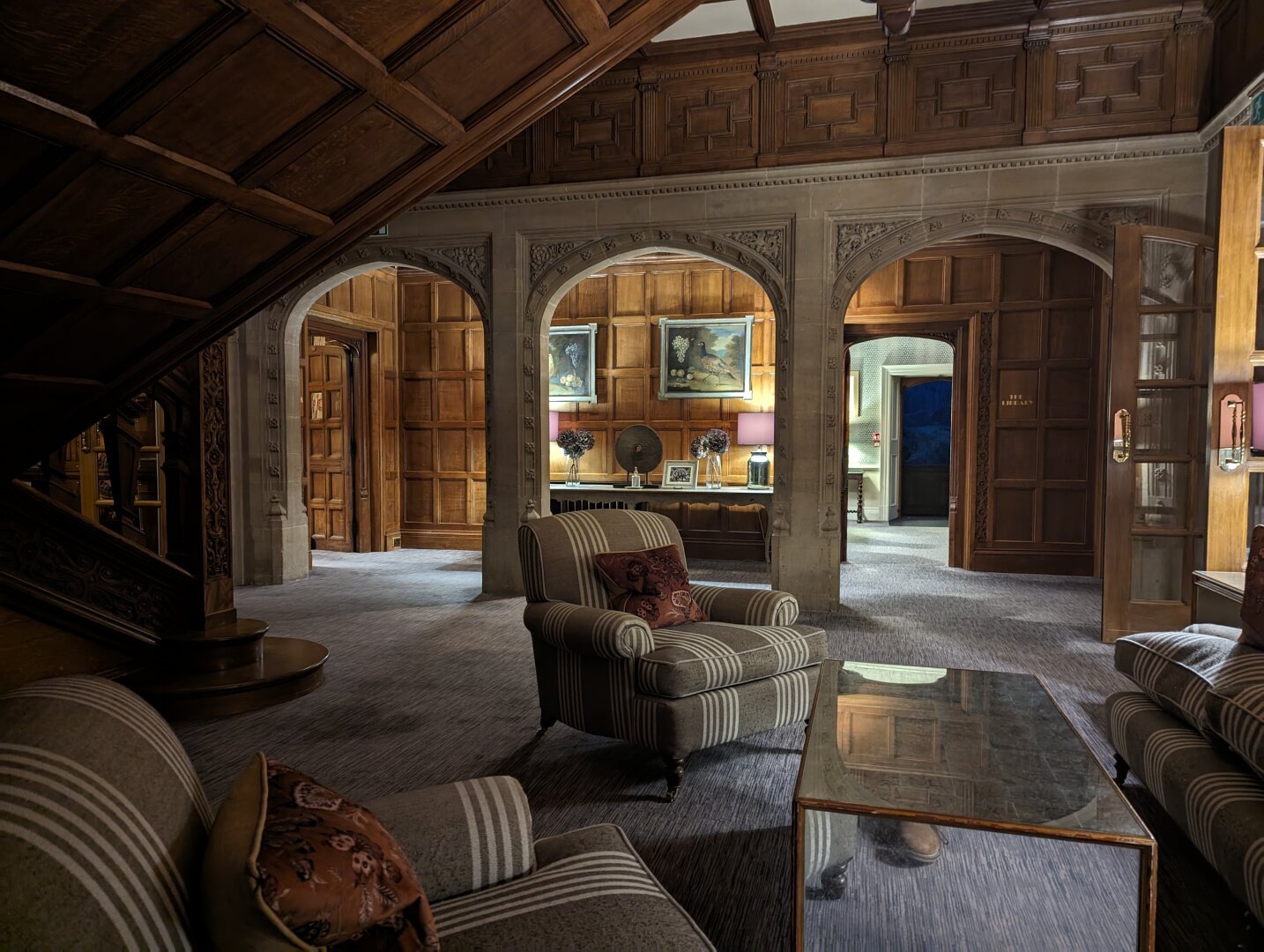 A dimly lit snug area under the stairs at Bovey Castle. There are archways in the back of the picture out into corridors. There are armchairs facing a fireplace in the foreground.