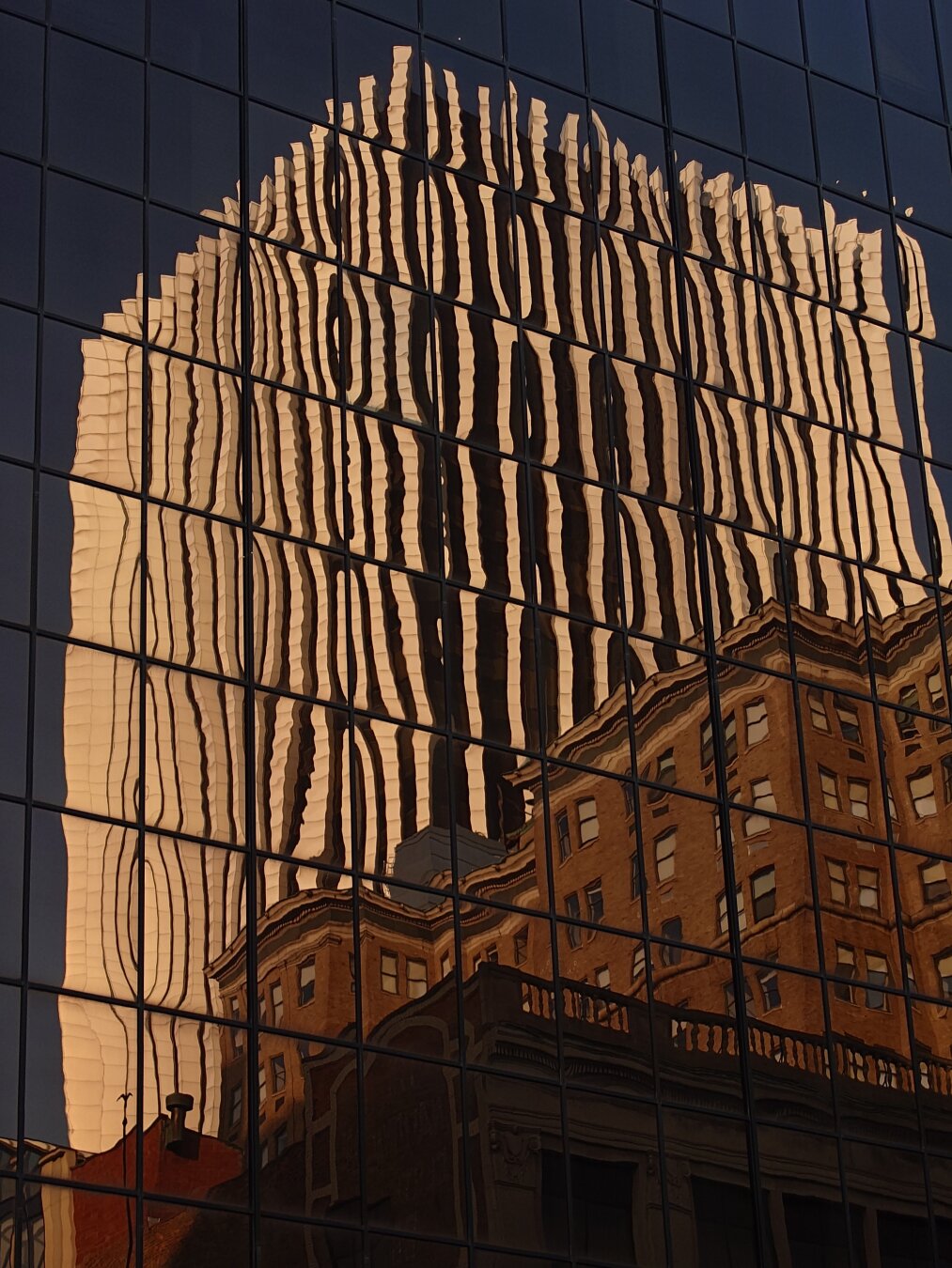 Photo of The Metropolitan tower, a glass skyscraper with white vertical ribs going up the height of the building, leering over old brick buildings and viewed in the reflection of another all glass building in the pale orange light of sunset.