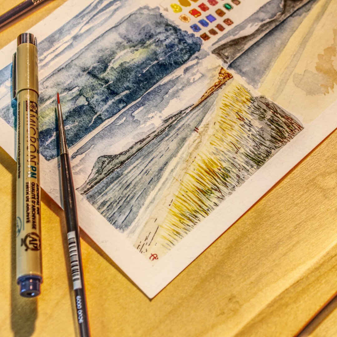Photo of a piece of watercolour paper on which a pencil and a brush are lying. In the postcard-sized painting, running from bottom left to centre right, you can see a dune with long, dry dune grass at sunset, with a beach at low tide behind it. A mountain range can be recognised on the horizon behind the sea. On the beach, two people can be recognised looking out to sea. Above and beside the painting, there are trials and colour tests on paper.