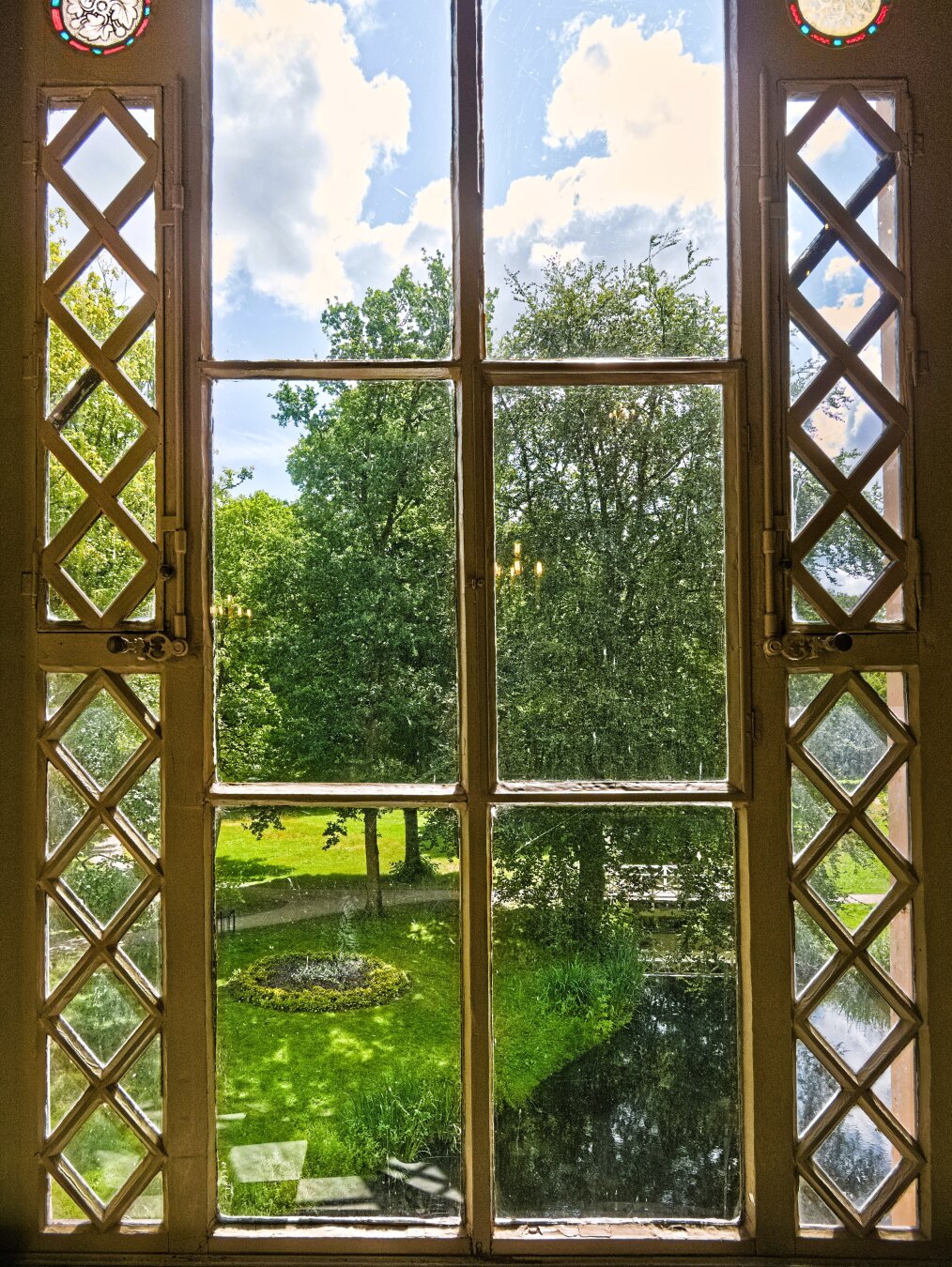 I look out of a window and see a park with green spaces, tall trees and a stream, spanned by a white wooden bridge. The window is very pretty. To the right and left, one above the other, are two narrow panes of glass, between whose brown wooden frames wooden chequered patterns can be seen. Between the narrow panes of glass – in the centre of the window – there are three panes of glass in each of two rows. Right at the top, above the narrow panes with the chequered patterns, there is a round window on each side. Its ‘frame’ is red with blue dots, and floral motifs can be seen inside.