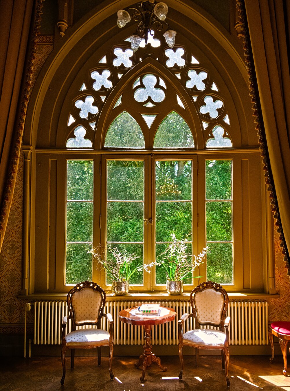 We are looking from inside at a Neo-Gothic-style pointed-arch window. The pointed-arch window is a large, magnificent wooden window (consisting of four panes side by side) with delicate tracery in the upper section. In front of it stands a seating arrangement: two classically upholstered armchairs with light-coloured upholstery. They flank a small, round wooden table. On the windowsill behind the armchairs stand two silver vases containing delicate white flowers. A radiator can be seen beneath the windowsill (not particularly Neo-Gothic ;)). The room features elegant wooden parquet flooring, patterned wallpaper and heavy curtains. Everything is kept in light colours. Sunlight streams through the window; outside the window, tall trees with lush foliage and the sky can be seen.
