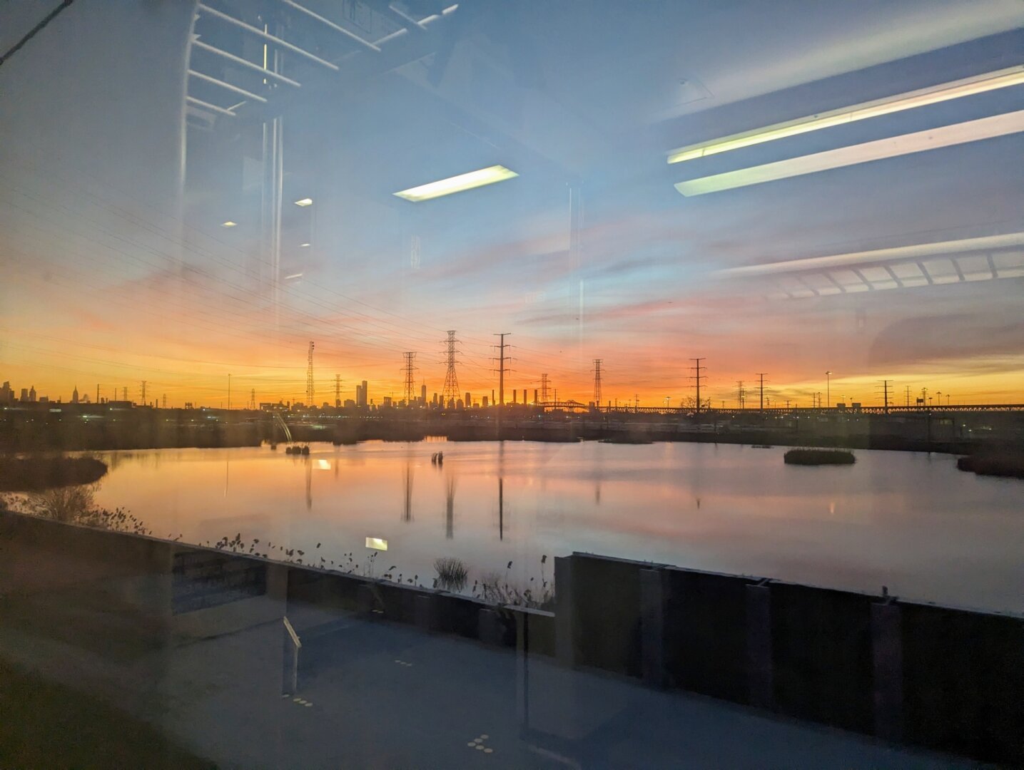 Fluorescent lights from inside the train car redirect off the window. The colors of the sunrise, orange and blue, reflect off of some body of water outside. Red clouds provide a backdrop for the distant New York City skyline.