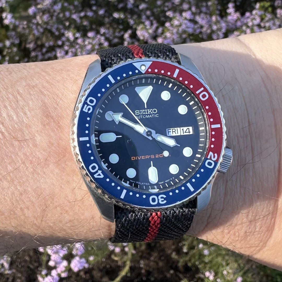 Watch with brushed stainless steel case with polished chamfered edges, dark blue dial with day and date at 3:00, inverted triangular index at 12:00, elongated indices at 6:00 and 9:00, circular indices elsewhere, crown at 4:00, dive bezel with blue and red aluminum insert, on a  navy blue woven nylon strap with red center stripe.