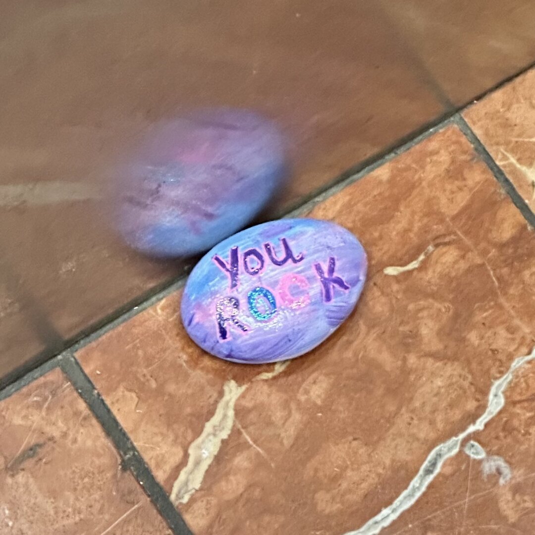 An image with caption: An oval-shaped stone painted in shades of blue and purple with “You ROCK” painted in purple letters outlined in pink. 