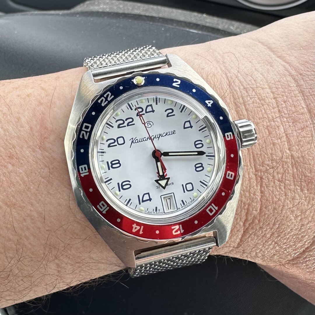 A wristwatch with a brushed octagonal stainless steel case, a white 24-hour dial with dark blue numeric indices, arrow hands, a 6:00 date, and a red and blue 24-hour bezel, on a milanese mesh strap.