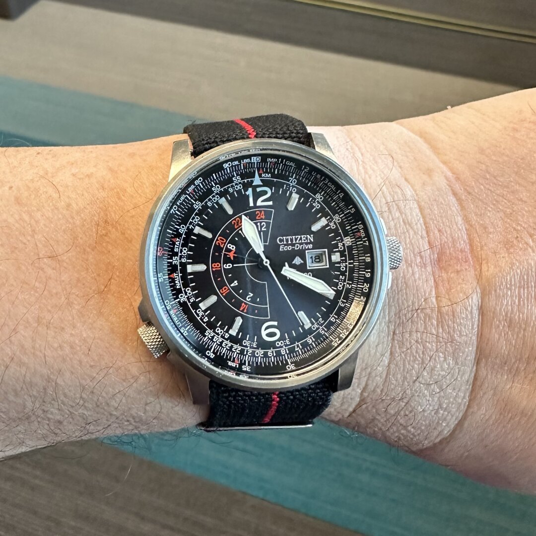 Watch with a black sunburst dial, white sword hands, numeric indices at 12 and 6, reflective faceted indices elsewhere, 8:00 crown that controls the slide rule bezel, GMT hand with one red and one white airplane-shaped hand for the 180° GMT scale, and date window at 3:00, on a black elastic fabric strap with red stripe.