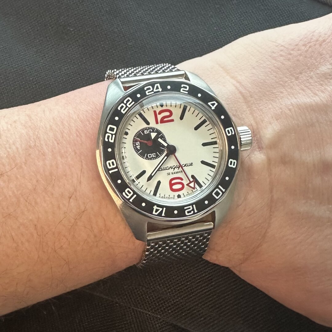 An image with caption: A watch with white dial, black seconds subdial at 10:00, red numeric indices at 12 and 6, black baton indices elsewhere, brushed stainless steel case, black 24-hour bezel with white hours, and a red GMT hand, on a milanese mesh bracelet.