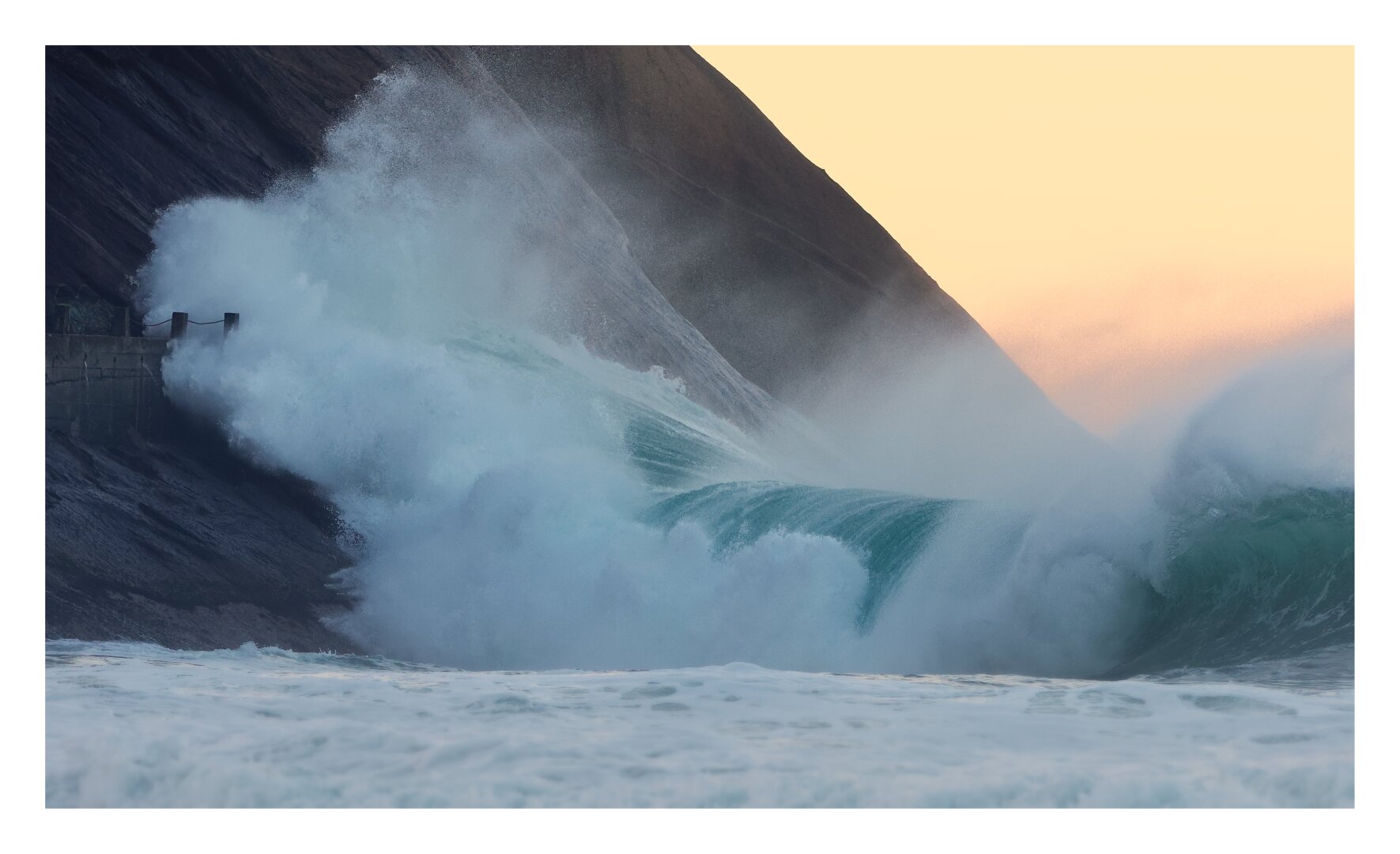 A huge turquoise wave crashes against a dark, steep mountainside, sending a huge whitewater and  spray into the air. There is a tiny surfer duckdiving the wave. The golden sky in the background contrasts with the cool blue tones of the turbulent ocean.