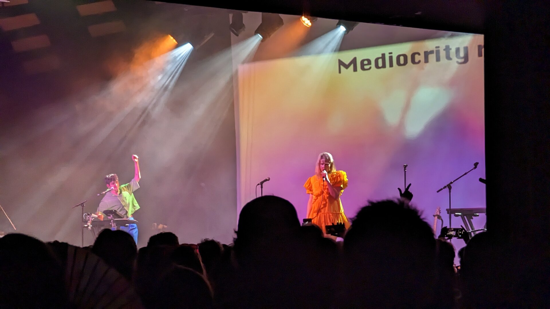 A photo of two members from the band La Tigre at a gig venue. Person on the left is playing synth and person on the right is singing. 

Silhouettes of audience heads can be seen at the bottom.

In the background the word "mediocrity" is projected onto a screen.