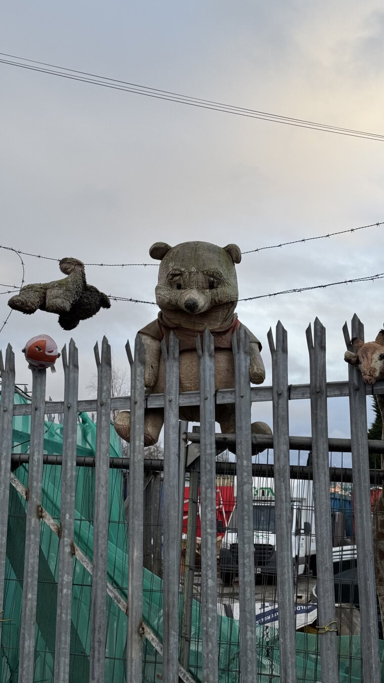 Centre of the photo has a worn Winnie the Pooh teddy, attached to a metal fence. There are other teddies to the left of the photo. In the background is a cloudy sky.