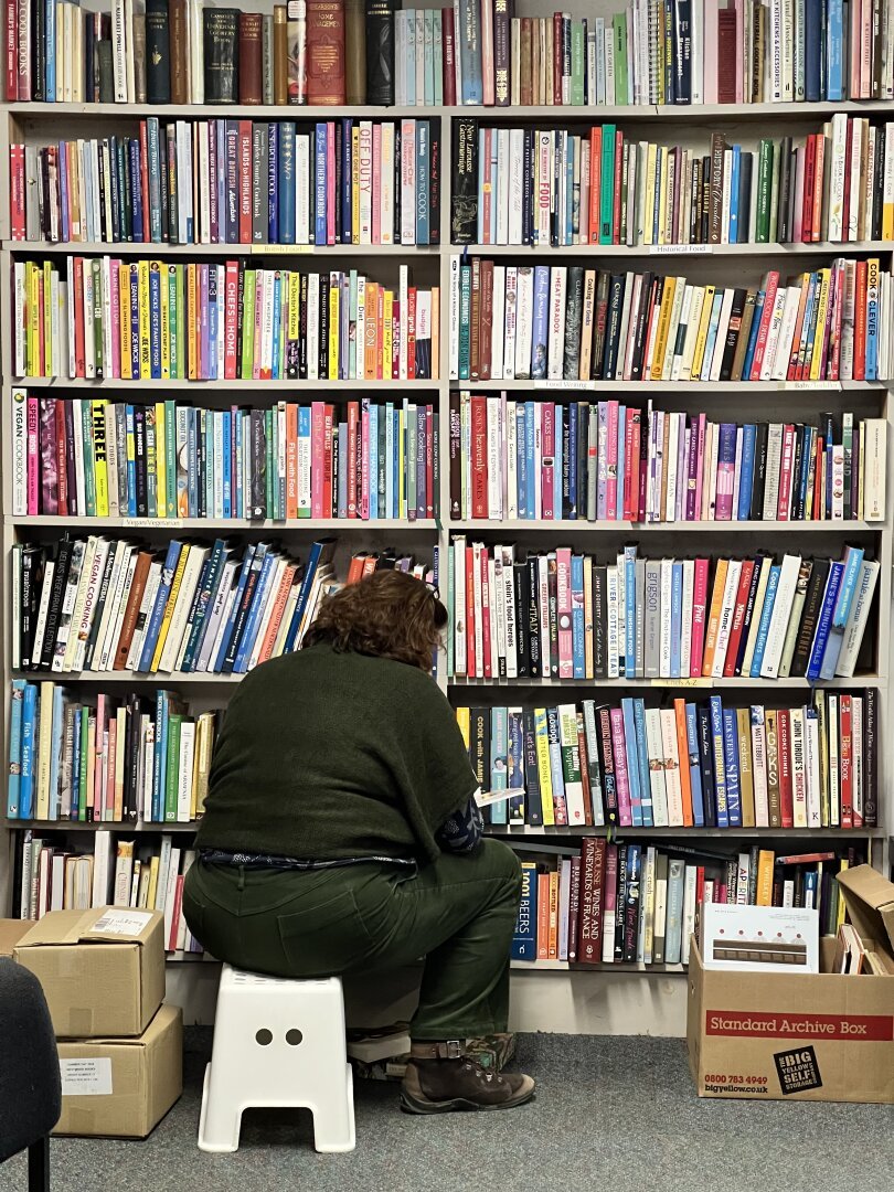 A person, dressed in dark green,    sitting on a low stool in front of crowded bookshelves in a second hand book shop in Sedbergh, UK