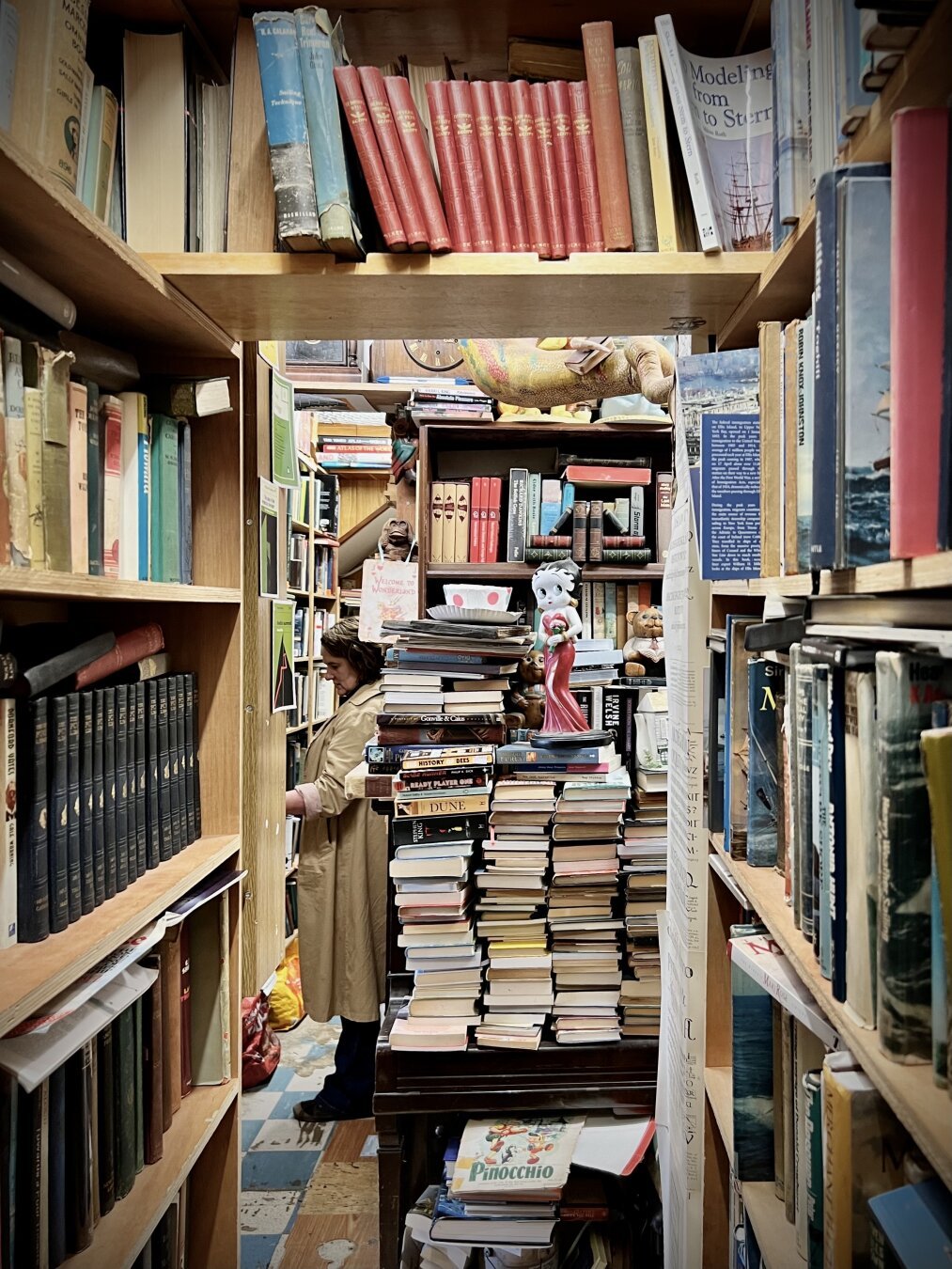 View through bookshelves laden with second-hand books to a door-like opening under another bookshelf, past some piles of books (spines not showing) to some bookshelves and a person reading a book, with bookshelves and stacked books behind them. No windows.