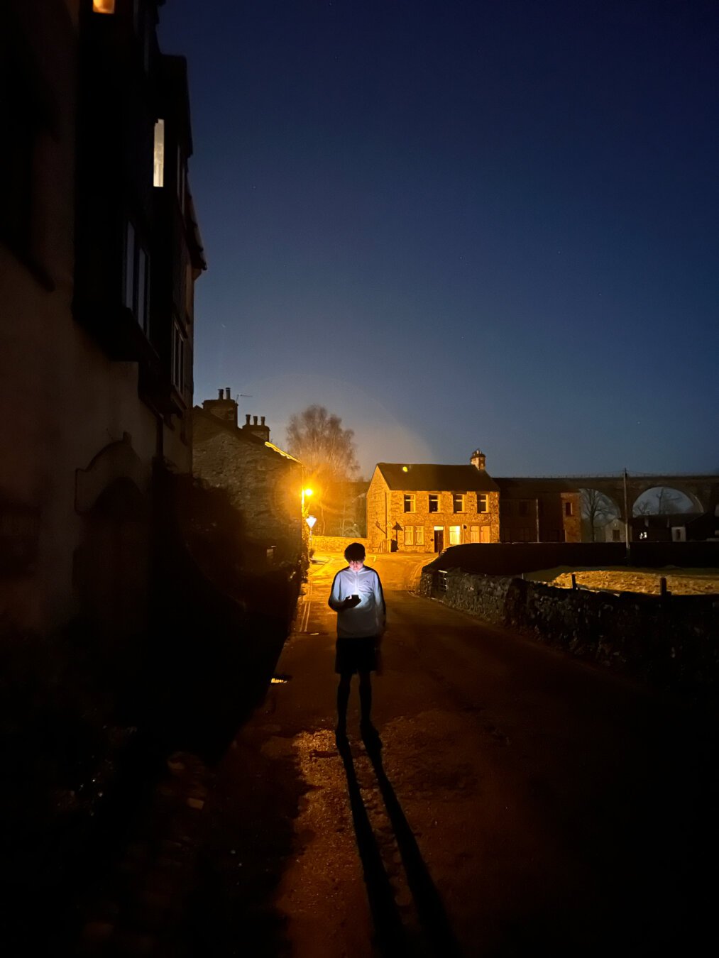 Person checking their phone at dusk, their face and upper body (dressed in a white track suit top) brightly illuminated by the white light of the screen, while the background is stone-built houses behind and left, the arches of a viaduct to the right, a dark blue gradient of a sky, and vivid yellow street and house lighting contrasting with the white of the phone