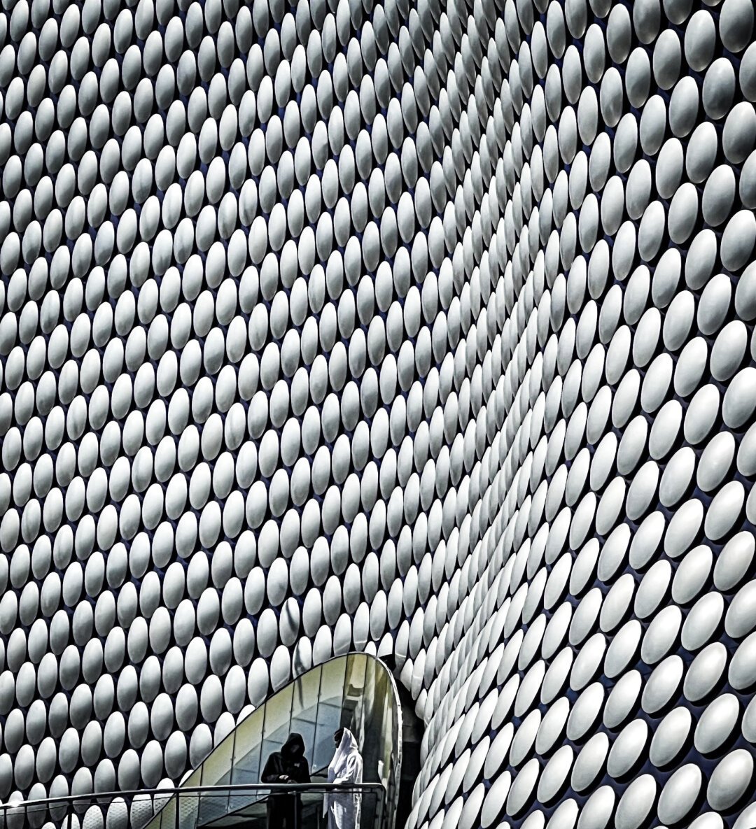 Two figures, one dressed in a hooded white robe-like garment, the other in black, stand in conversation at a rail on a balcony high above the viewer, framed in an arched entrance to a building that rises above them, clad in waves of large silver discs.

Ok… two people talking outside a shopping centre in Birmingham, UK