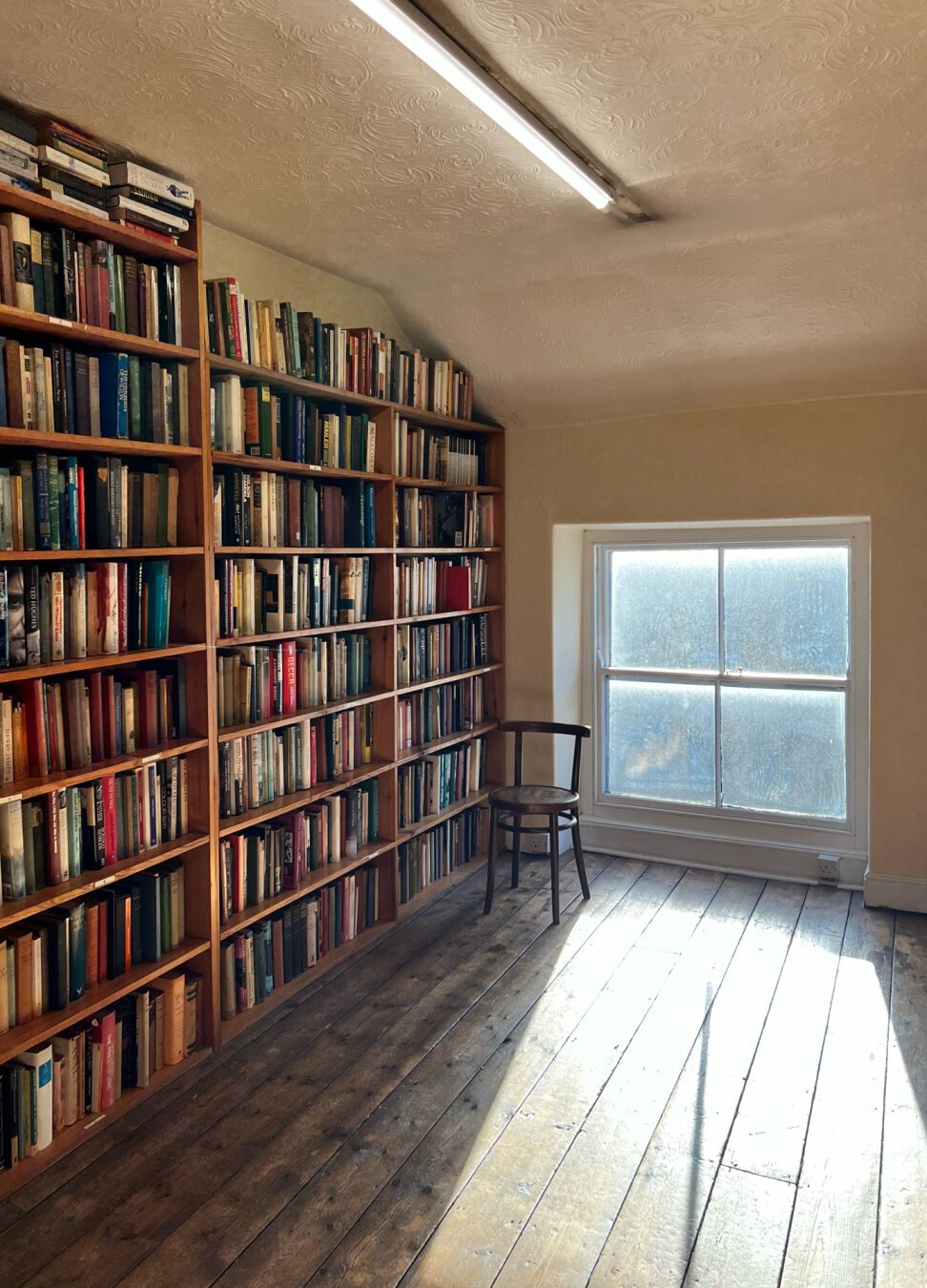 View from the doorway of a room on the third floor in a beautiful second hand bookshop in Carnforth, Lancashire (UK), with bookshelves filling the left wall, a low window casting light onto wooden floorboards in the foreground, and an old wooden chair next to the window for people to sit down and read. In the ceiling, a fluorescent tube light creates a diagonal.