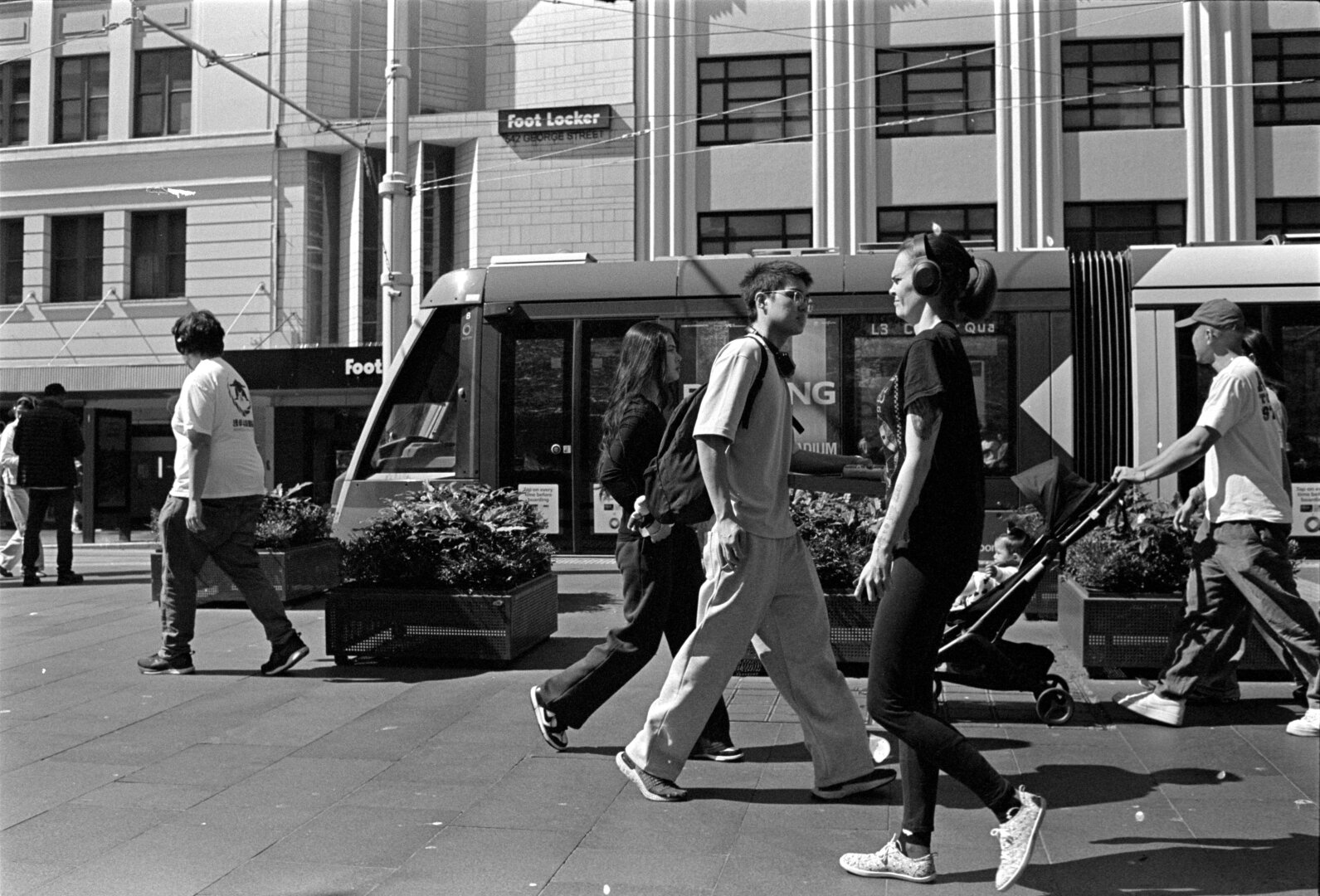 A B&W photo of people walking along the pedestrianised George St, in Sydney, passing a light rail train.