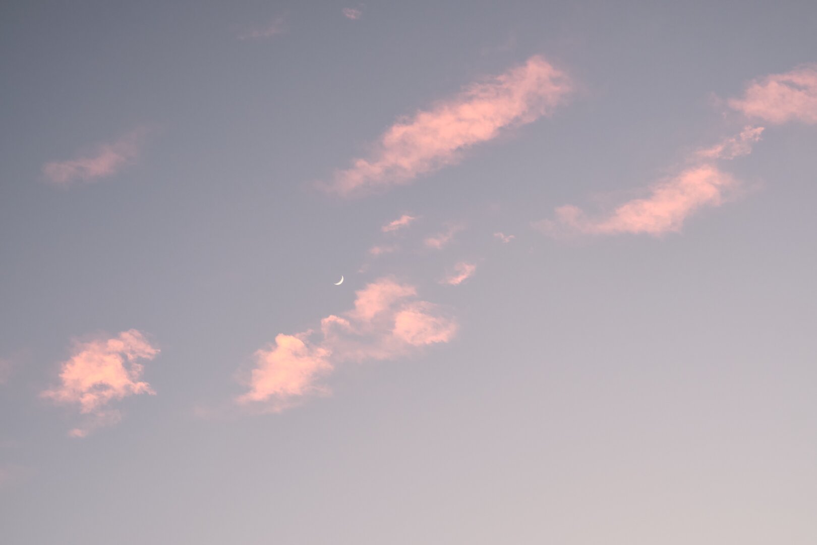 Photo of wispy pinkish clouds in a early evening blue hour sky with a small sliver of the moon visible.