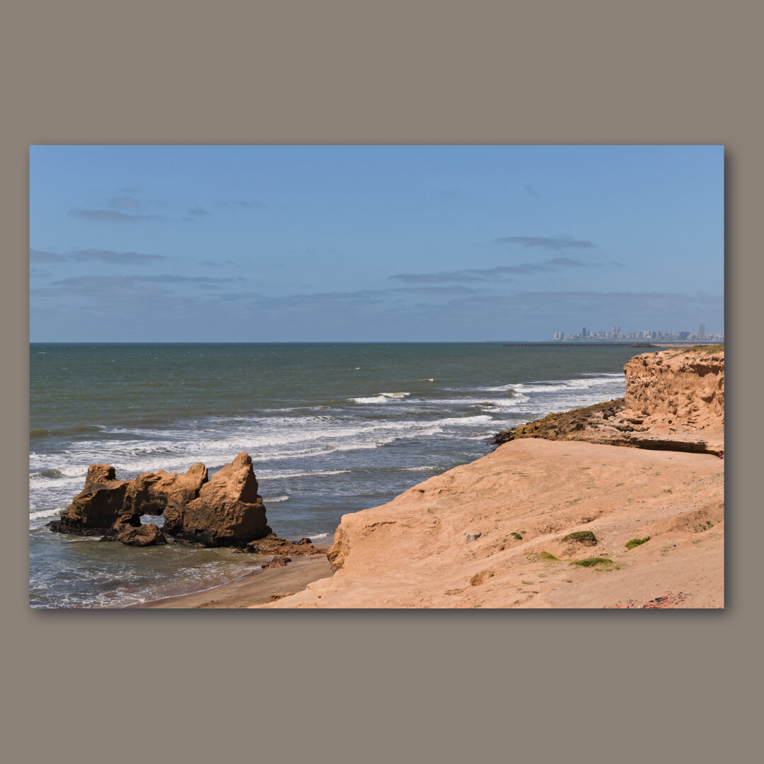 A cliff in front of the sea with a city behind.