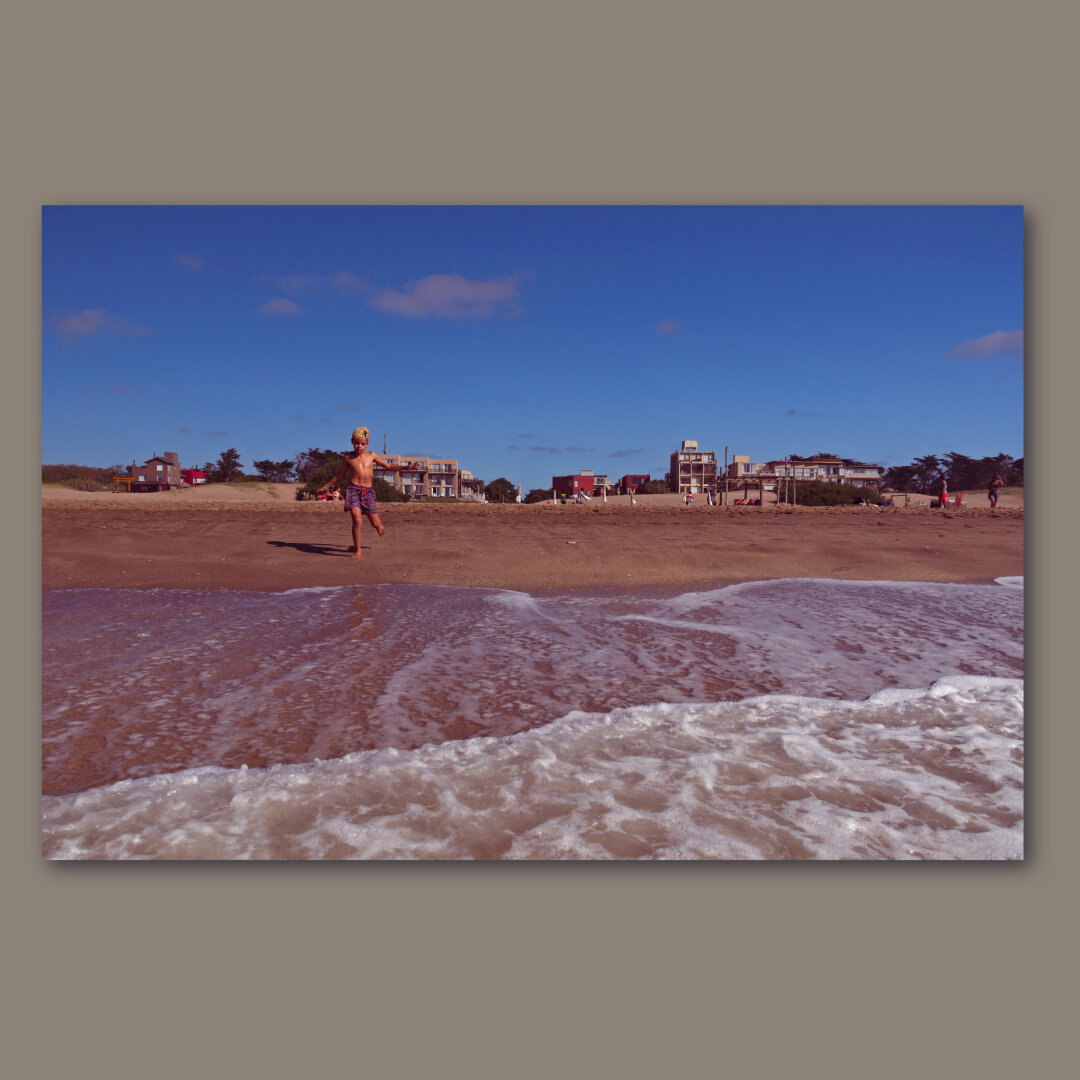 A boy running into the sea with some buildings far away behind him.