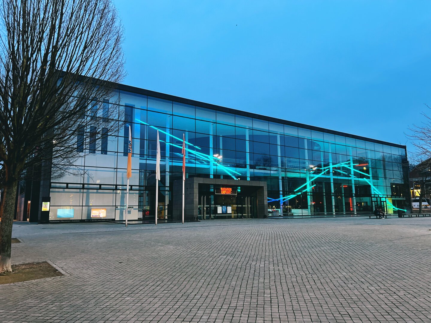 Zu sehen ist die Vorderseite des Erfurter Theaters. Hinter der Glasfassade zeichnet sich eine neonblaue Welle ab.