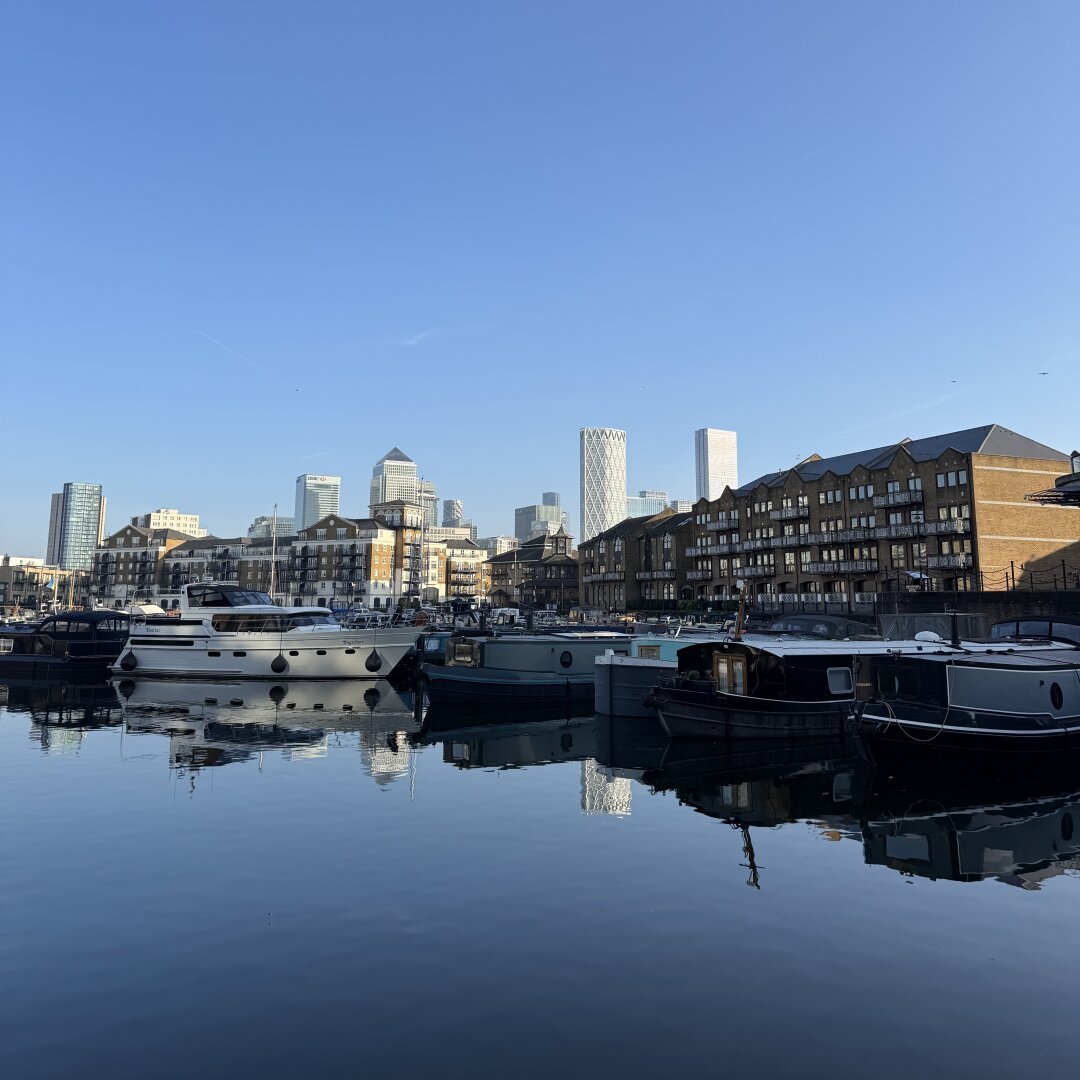 A photo of Limehouse marina in East London. Blue skies above are mirror in the calm water of the marina. Some houseboats in the foreground are dwarfed by the high-rise towers of Canary Wharf in the distance.