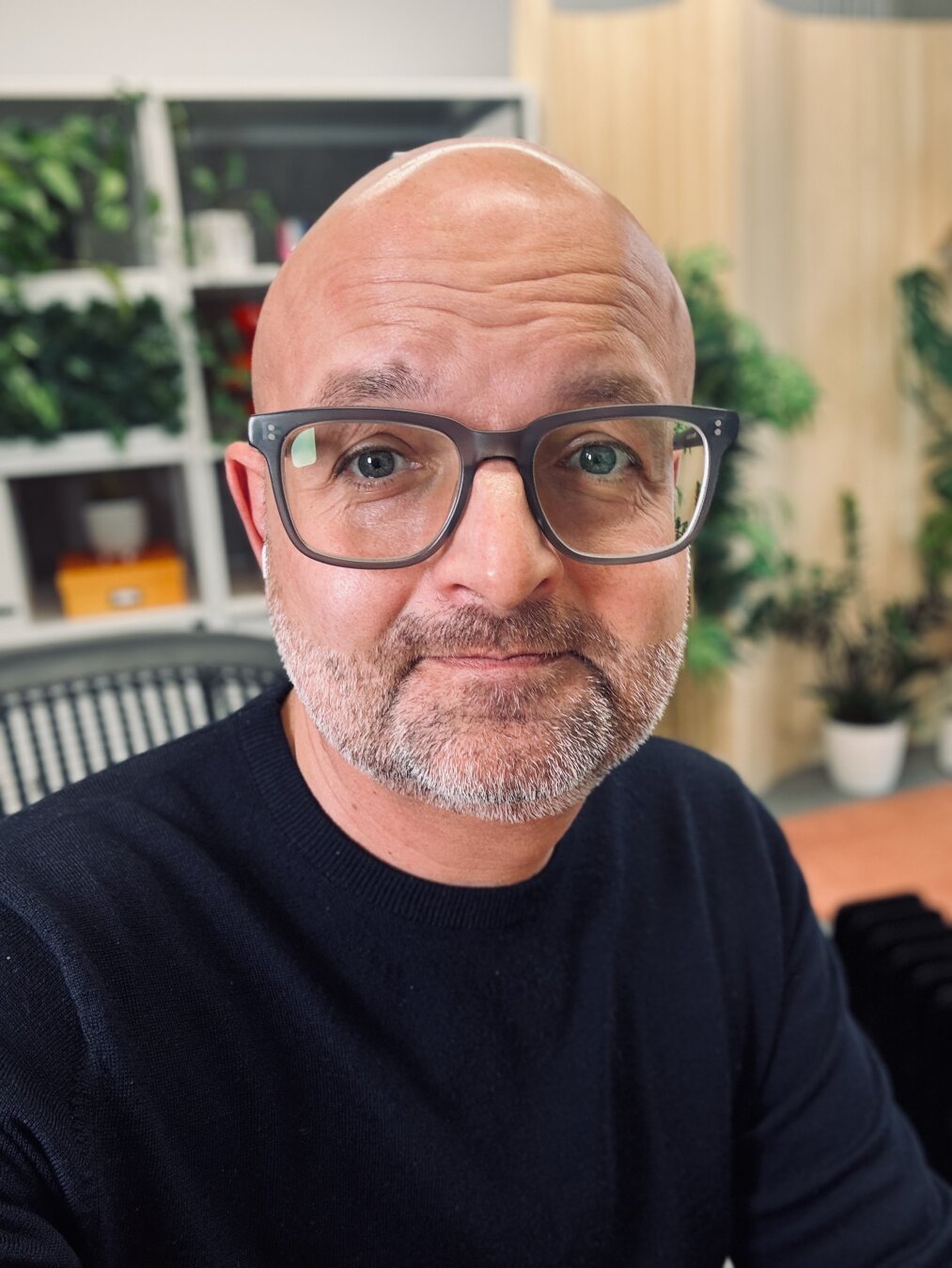 Selfie of a middle-aged bald, bearded man wearing glasses - which only slightly cover the significant bags beneath his eyes. He looks and feels tired. The photo was taken in his office, with lots of plants arranged on shelves in the background.