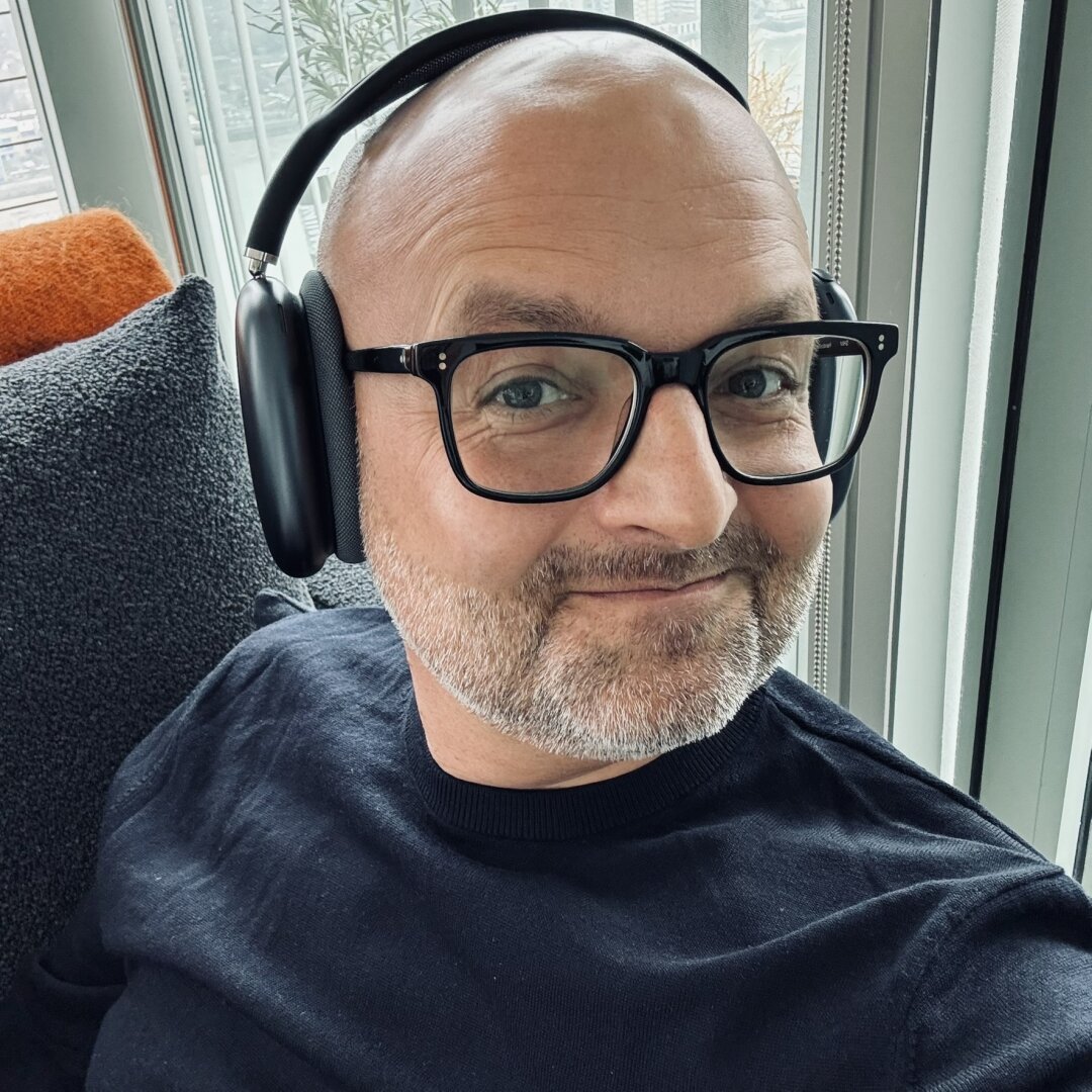 A selfie of a man wearing new Apple AirPods Max headphones. He's wearing a navy sweater, black-rimmed glasses, and a smile.