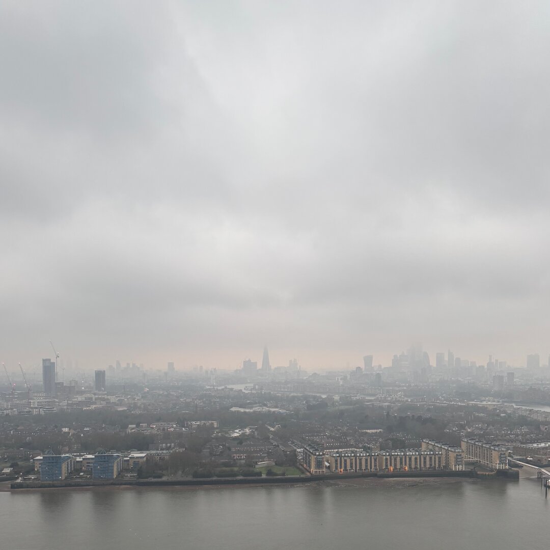 A pic of London’s skyline, looking west. Grey skies and clouds obscure the horizon. Even the Thames in the foreground looks a muddy grey colour.