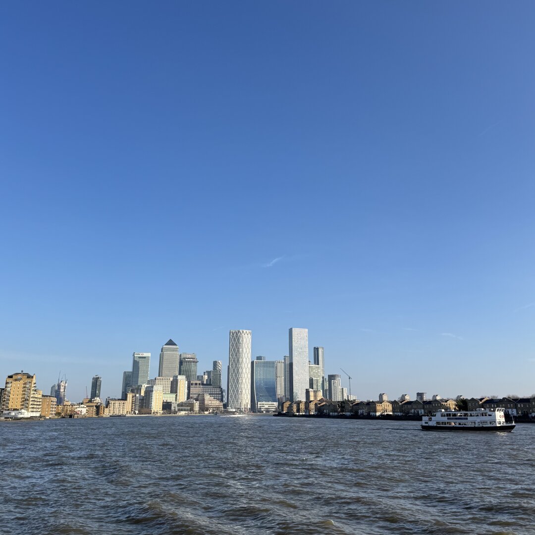 A photo of the Canary Wharf skyline, consisting of several large skyscrapers, taken from further along the River Thames in Wapping, East London. The skyscrapers are framed by bright blue skies and the slightly choppy River Thames in the foreground.