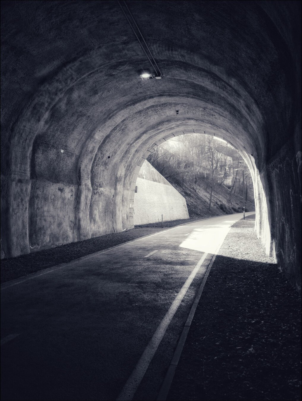 The photo depicts the interior of a dark, curved tunnel, with the road inside illuminated by soft, diffused light streaming in from the tunnel's open end. The tunnel walls are rough and textured, with visible wear, giving it an aged, slightly industrial feel. A single light fixture is mounted on the ceiling of the tunnel, contributing minimal brightness. Beyond the tunnel's exit, the outside environment is bathed in daylight, revealing trees, a slope, and faint outlines of structures, creating a strong contrast between the shadowy interior and the brightness outside. The perspective draws attention to the light at the end of the tunnel, symbolizing openness or escape.