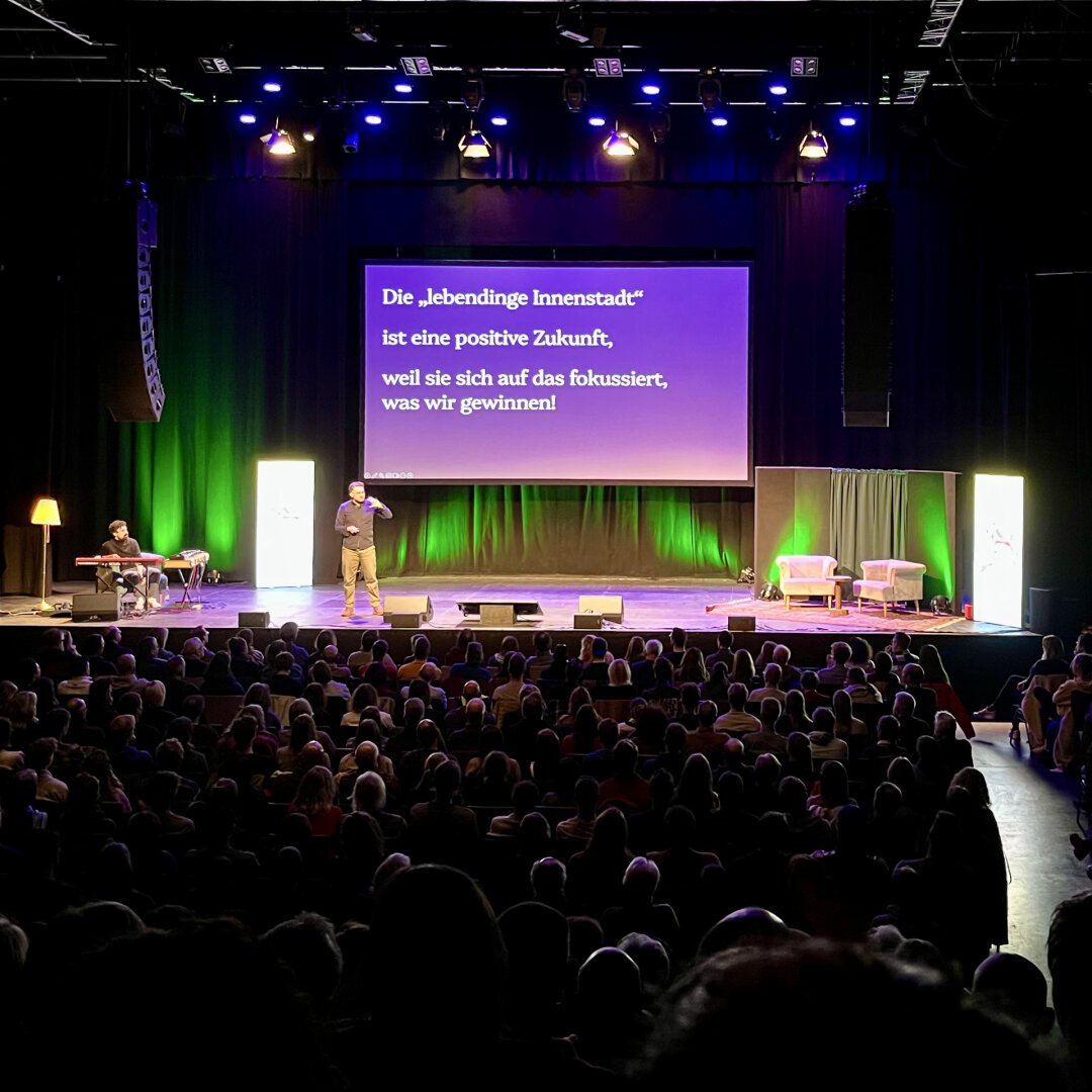 Johannes Kleske auf der Bühne des Science Slam im Tollhaus Karlsruhe. Auf der Folie seiner Präsentation steht weiß auf lila Hintergrund: »Die „lebendige Innenstadt“ ist eine positive Zukunft, weil sie sich auf das fokussiert, was wir gewinnen!«