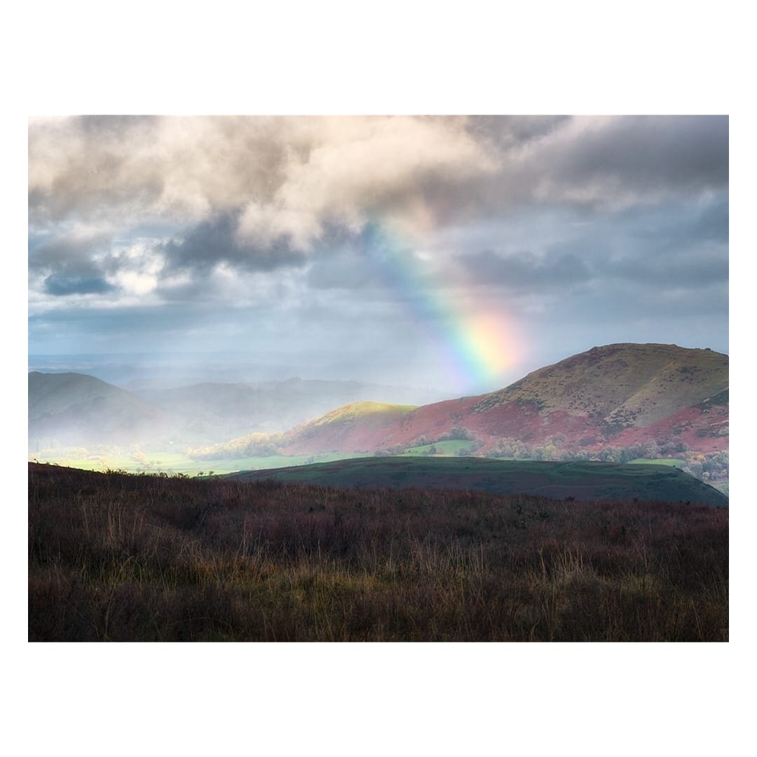A dramatic rainbow arcs across a cloudy sky above a sweeping autumnal landscape, with the foreground composed of dark reddish-brown moorland grasses and low heather in the foreground, whilst distant rounded hills rise in layers of russet, mauve, and sage green beneath bands of mist and atmospheric haze. The middle distance reveals pastoral fields in soft greens and golds nestled between the undulating slopes, with Caer Caradoc's distinctive rounded summit prominent on the right, its flanks painted in autumn hues of burgundy and burnt sienna. Brooding storm clouds dominate the upper half of the composition, their grey and white masses illuminated by breaks of pale blue sky through which the rainbow glows brilliantly, creating a moody yet uplifting vista characteristic of the Shropshire Hills' dramatic weather and rugged moorland terrain.