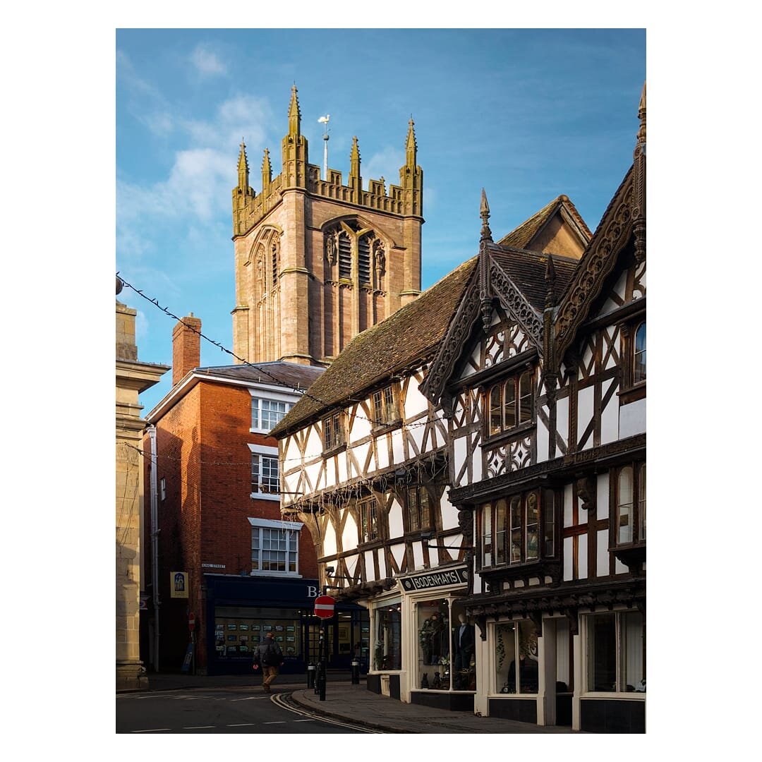 A picturesque street scene from Broad Street in Ludlow showcases a striking contrast between architectural periods, with a timber-framed building featuring distinctive dark wooden beams and white infill panels dominating the foreground on the right, its ornately carved bargeboards and leaded casement windows exemplifying Tudor craftsmanship. Behind and rising majestically above the period buildings stands St. Laurence's Church, its golden sandstone tower soaring skyward with multiple decorative pinnacles and a row of tall, pointed lancet windows that speak to its Perpendicular Gothic heritage. To the left, a later red-brick Georgian building provides contemporary context to the medieval street layout, whilst ground-floor shopfronts including what appears to be a clothing retailer occupy the street level, and a clear blue sky with scattered white clouds illuminates the honey-coloured stonework, creating a scene that encapsulates Ludlow's rich historical layers.