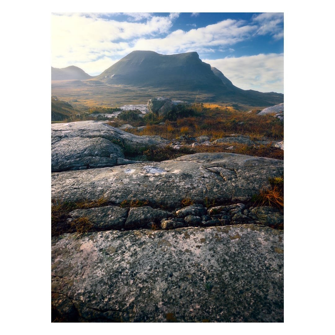 A portrait-format landscape photograph taken low to the ground shows the broad, flat-topped bulk of Sàil Gharbh, part of the Quinag massif in the north-west Highlands of Scotland, rising beyond an open expanse of moorland. In the foreground, large slabs of pale grey rock fill the lower half of the frame, their surfaces mottled with black, olive and ochre lichen and marked by cracks, small hollows and a few tiny pools of standing water. Tufts of rust-orange and golden grass and low heather grow in the fissures between the slabs and spread across the middle distance, where a few rounded boulders sit among the autumn vegetation and catch patches of warm sunlight. Beyond the moor, the mountain's smooth, shadowed slopes are dark green and blue-grey, with a steep drop falling away to the right and a lower rounded hill visible to the left, softened by a hazy glow. Above, a bright blue sky is streaked with wispy white cloud, and strong backlight from the left of the frame gives the whole scene