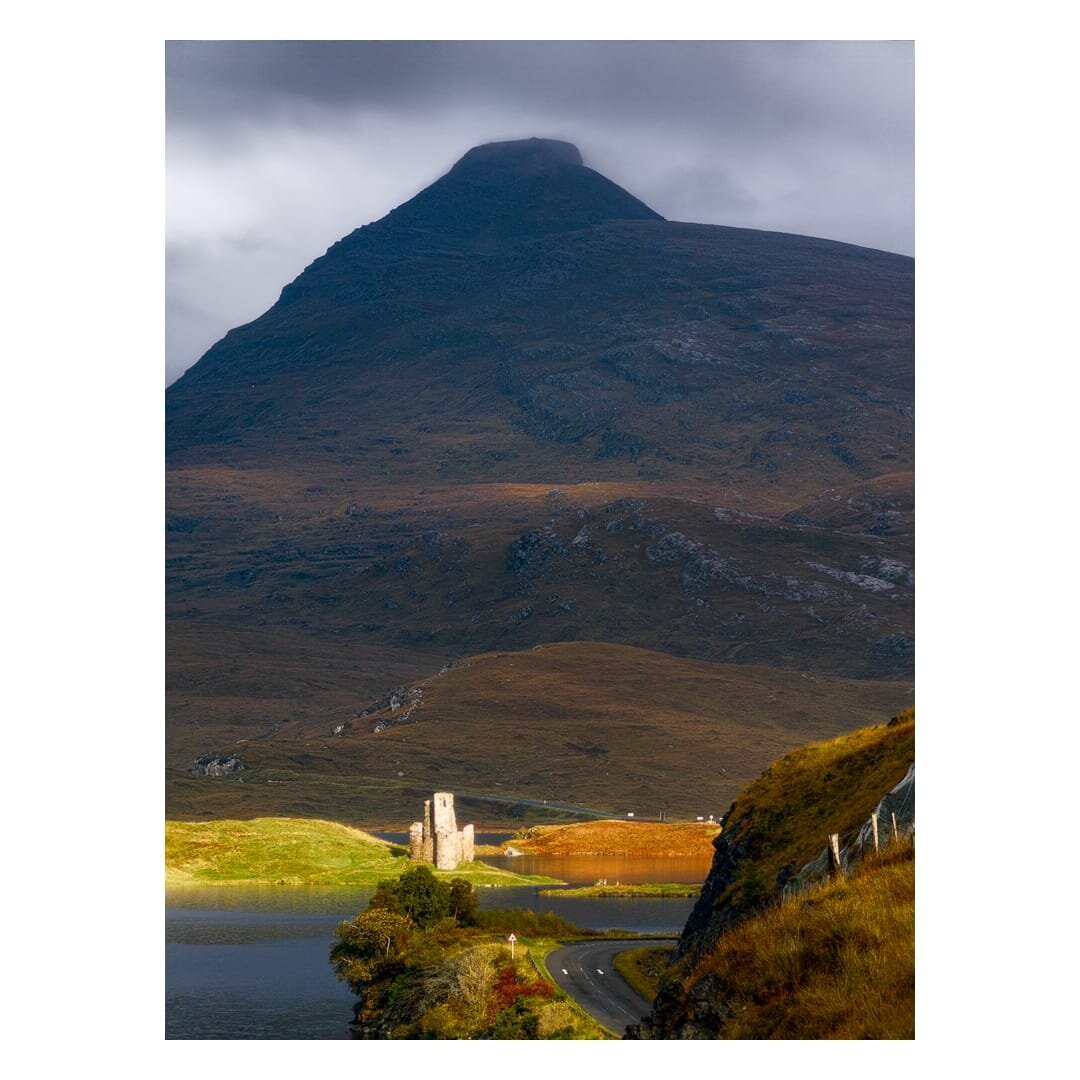 A portrait-format landscape photograph taken in autumn light shows the ruins of Ardvreck Castle on a small green promontory jutting into a dark loch, with the great bulk of Quinag rising above in the Scottish Highlands. A narrow, pale stone tower and low broken walls stand out brightly against the moody scene, lit by a shaft of sunlight that picks out the grass in vivid yellow-green and turns a stretch of water behind the castle a glowing copper-orange. Above, the huge mountain fills most of the frame, its steep, shadowed slopes in deep brown and charcoal with scattered grey outcrops of rock, and its flat-topped summit is partly wrapped in heavy slate-grey cloud. In the lower right, a bank of rusty-gold bracken and grass slopes down beside a dark crag, with a rock-netted fence and wooden posts, while a single-track road curves away from the foreground past a small triangular warning sign and a cluster of trees in russet and yellow autumn colours. The overall mood is dramatic and broodi