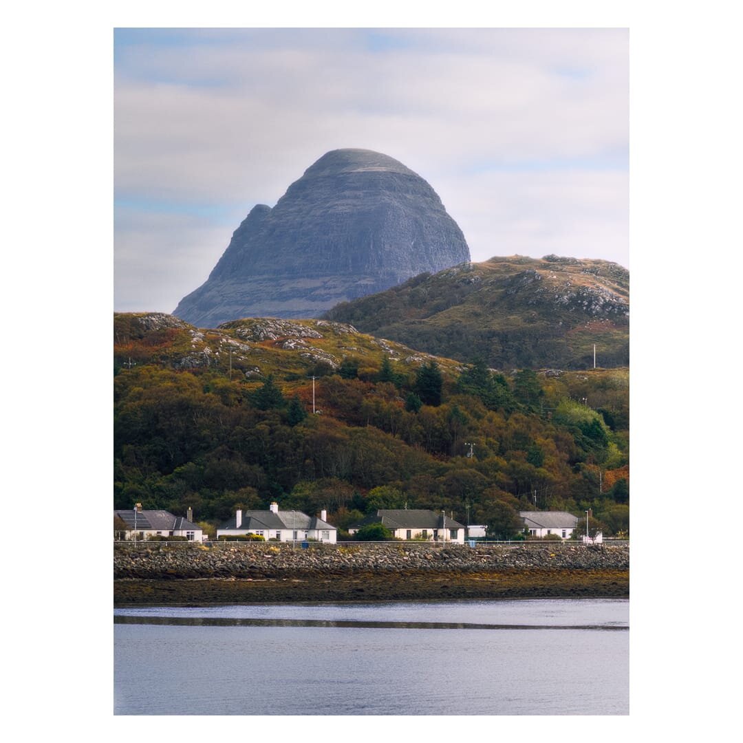 A portrait-format landscape photograph shows the distinctive dome of Suilven rising above the village of Lochinver in the north-west Highlands of Scotland, seen across a calm stretch of grey-blue loch in the foreground. The mountain's smooth, rounded summit and steep flanks are banded with layers of dark grey sandstone, and a smaller craggy shoulder juts out to its left against a pale sky of soft white and blue-tinged cloud. Below the peak, rolling hills rough with grey rock outcrops and tawny moorland grass lead down to a thick, wooded slope in rich autumn shades of olive green, rust, ochre and deep brown, scattered with dark conifers and a few telegraph poles. Along the shoreline at the bottom of the hillside sits a row of low, white-painted cottages with slate-grey roofs and chimneys, separated from the water by a low stone sea wall and a broad band of rocky, seaweed-covered shore in muted gold and brown. The mood is quiet and still, with the muted light giving the scene a calm, und