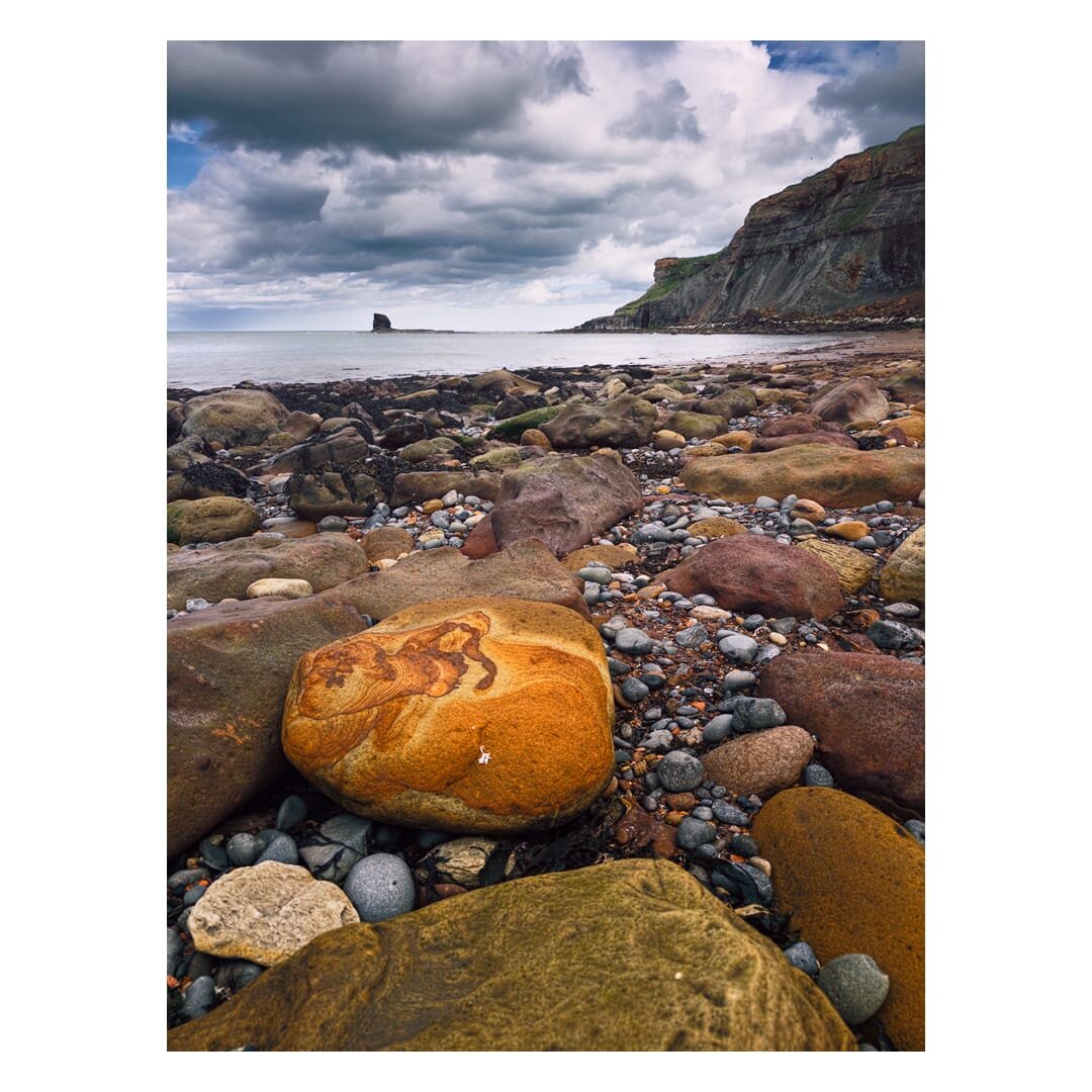 A boulder-strewn shoreline in Saltwick Bay, viewed from the main central beach area looking towards the southeastern cliffs and Black Nab. In the foreground, a striking rounded sandstone boulder shows bold swirling patterns in burnt orange and rust, its surface weathered into flowing bands quite different from the duller mudstone and pebbles surrounding it, which range from grey and buff to deep plum and olive. Beyond this scatter of rocks and shingle, a calm grey sea stretches to the horizon, where the dark pinnacle of Black Nab is just visible silhouetted against the pale water. To the right, the layered sandstone and shale cliffs curve away towards the headland, their strata picked out in ochre and green where vegetation clings to the upper ledges, while overhead a heavy bank of grey cloud dominates the sky, with only a small patch of blue breaking through to the left.