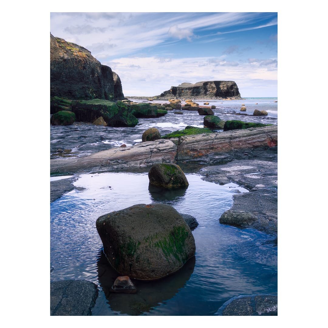 A low-tide seascape at Saltwick Bay, taken from beneath the northeastern cliffs and looking out towards Saltwick Nab. In the foreground, calm rock pools reflect a pale blue sky streaked with wispy cloud, with several large, rounded boulders draped in dark green seaweed rising from the still water. Flat, weathered sedimentary rock ledges stretch across the middle of the scene, their surfaces banded in grey and rust-red tones and pitted with smaller pools. To the left, a dark, jagged cliff face rises steeply, its upper ledges tinged with moss and lichen, while further along the shoreline scattered boulders lead the eye towards the distant flat-topped headland of Saltwick Nab, its layered rock strata clearly visible against the sea beyond. The overall mood is quiet and atmospheric, with soft natural light and gentle reflections capturing the exposed, sculptural character of this stretch of Yorkshire coastline at low tide.