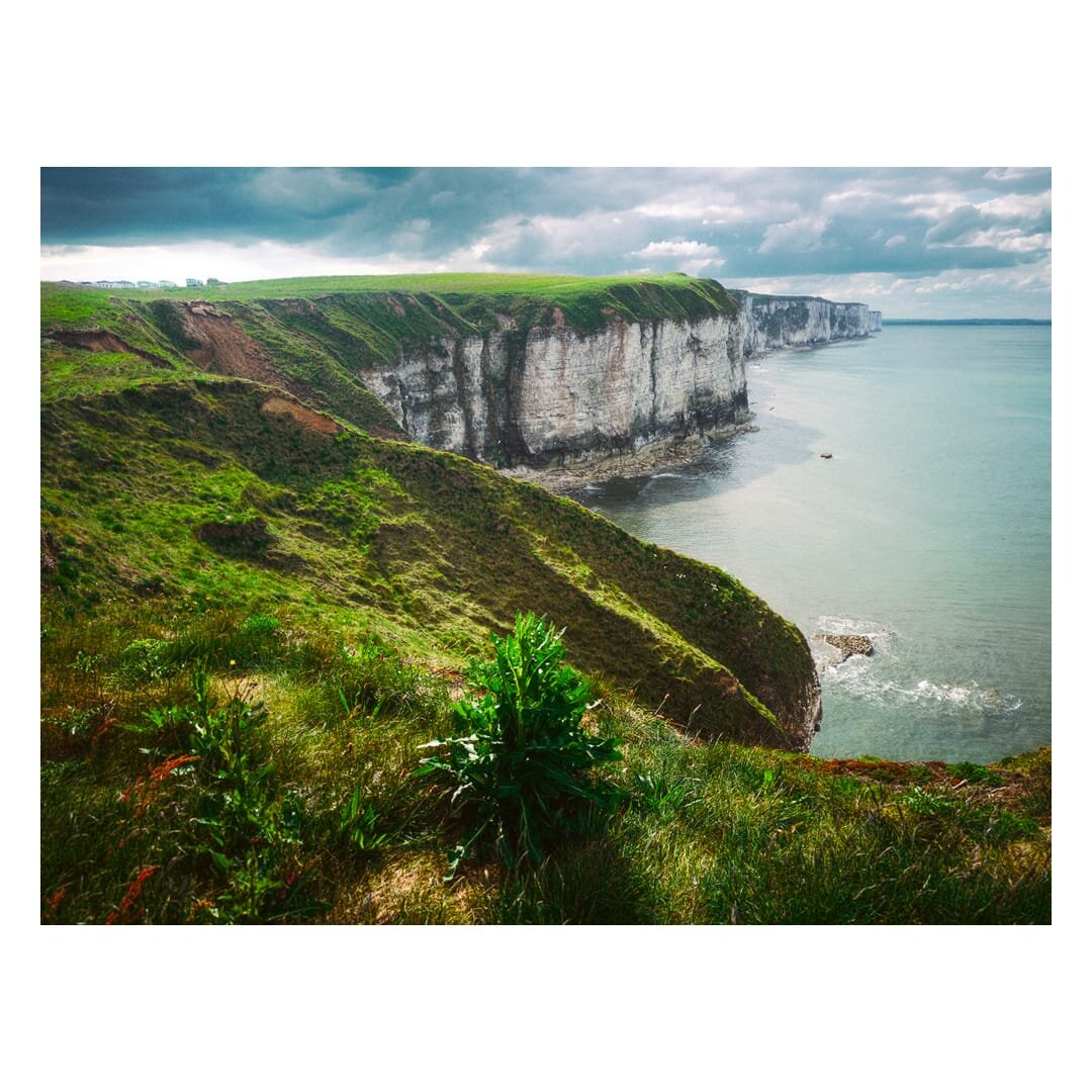 View west along the Bempton Cliffs, with towering white chalk faces topped by vivid green grass stretching into the distance beside a calm grey-blue sea, beneath a dramatic sky of dark and broken clouds.