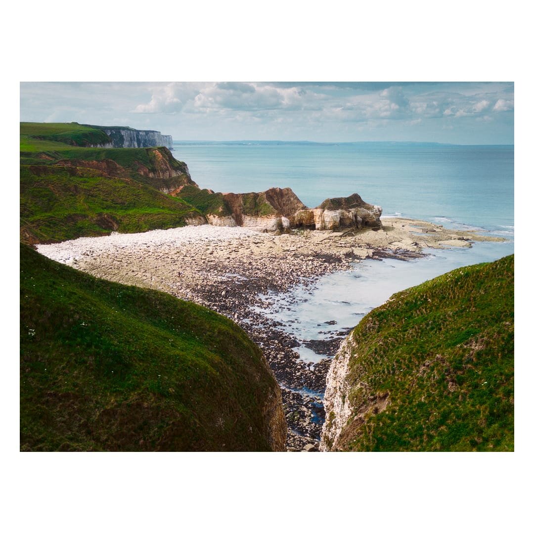 View from the cliffs overlooking Thornwick Bay on the Flamborough coastline in East Riding of Yorkshire, with steep green headlands surrounding a broad rocky beach and pale chalk formations jutting into the calm blue sea beneath a partly cloudy sky.