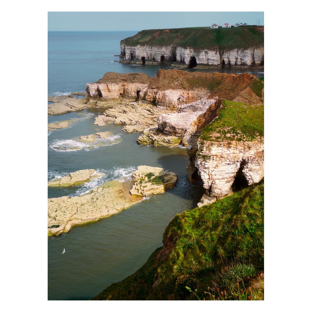 Clifftop view west of Thornwick Bay looking east across the rugged Flamborough coastline, with steep grass-topped chalk cliffs, sea caves and pale rocky platforms extending into calm blue-grey water, and a few houses visible on the distant headland.
