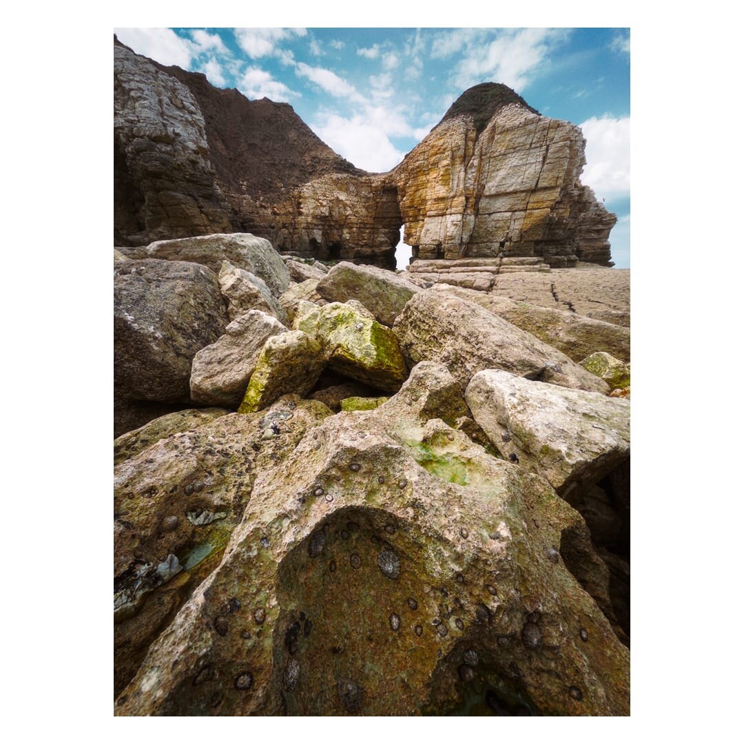 Low-angle view across large, weathered rocks at Thornwick Bay towards Thornwick Nab, where layered chalk cliffs rise steeply beneath a blue, partly cloudy sky, with a narrow natural opening visible through the rock formation.