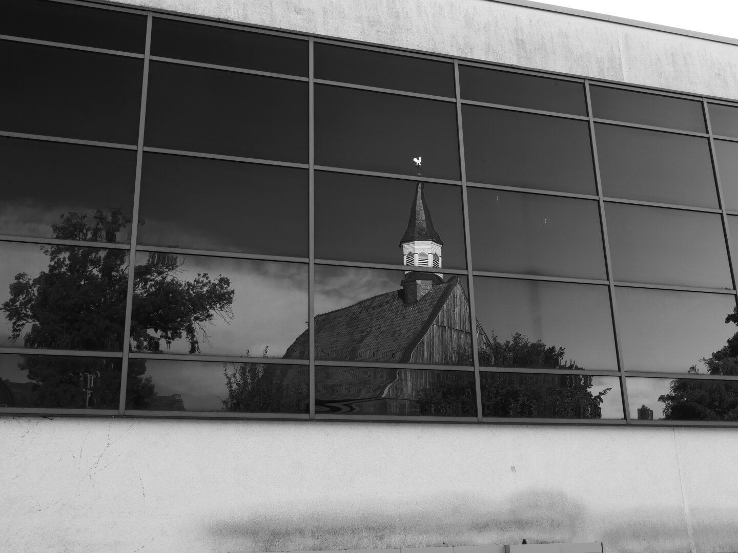 Schwarzweißaufnahme einer modernen Glasfassade. In den dunklen Fensterscheiben spiegelt sich eine ältere Kirche (Alte ev. Kirche in Wattenscheid) mit steilem Dach und kleinem Turm sowie umliegenden Bäumen und Wolken. Das regelmäßige Raster der Fassade kontrastiert mit der historischen Architektur im Spiegelbild.