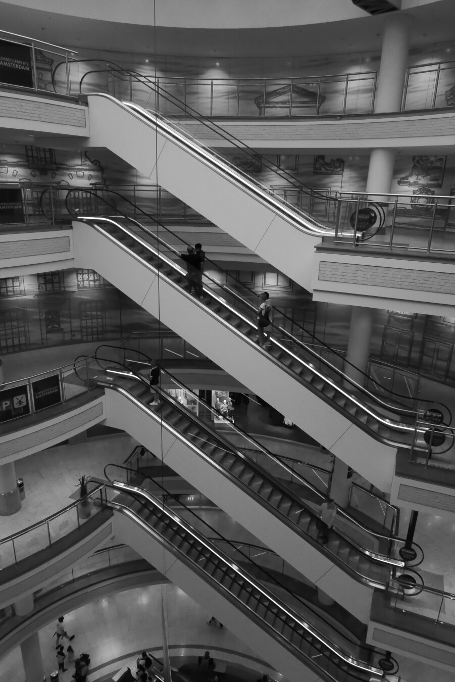 Schwarzweißaufnahme des mehrgeschossigen Innenraums des Einkaufszentrums Limbecker Platz in Essen.
Mehrere Rolltreppen kreuzen sich diagonal über verschiedene Ebenen und bilden ein dichtes geometrisches Muster aus Linien, Geländern und Brüstungen. Einzelne Menschen fahren auf den Rolltreppen oder bewegen sich auf den Etagen darunter.